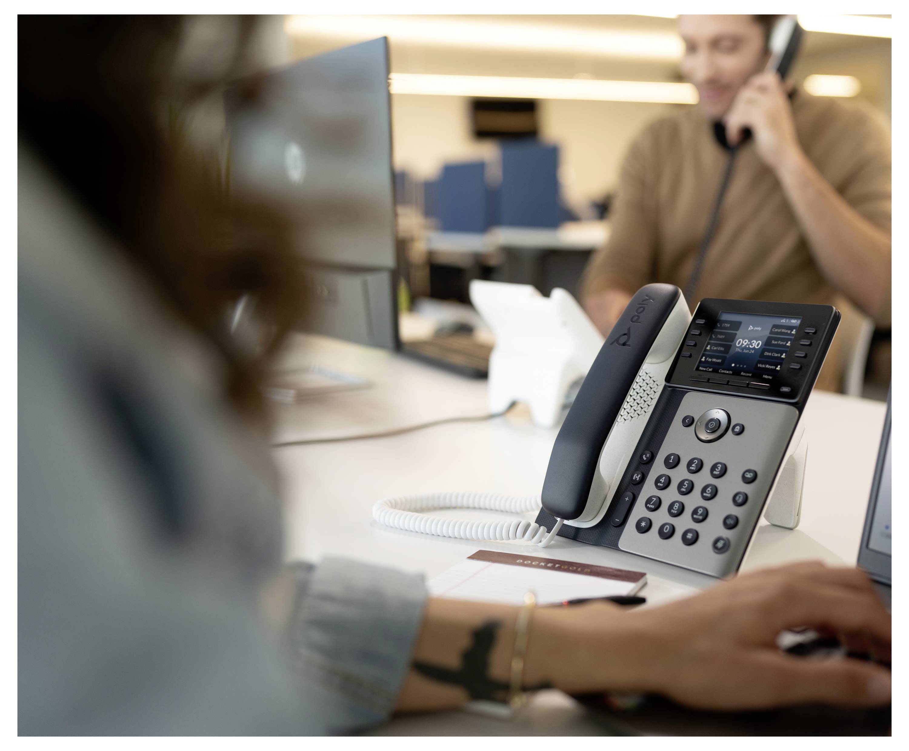 Bürotisch mit einem Schreibtischtelefon im Vordergrund und zwei Personen, die im Hintergrund arbeiten, eine spricht telefoniert.