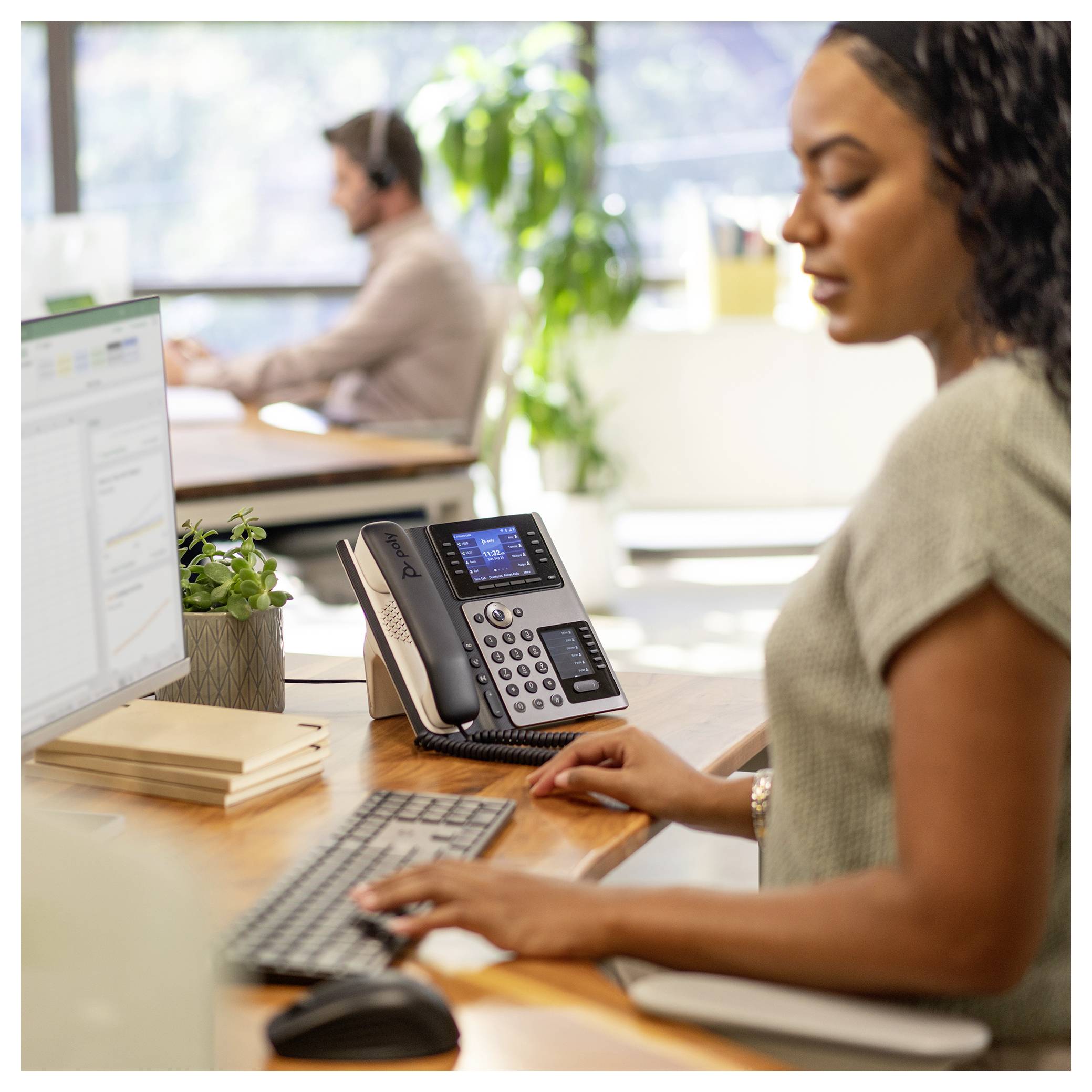 Frau tippt an einem Schreibtisch in einem Büro, nutzt einen Computer und ein Schreibtischtelefon; Kollege arbeitet im Hintergrund.