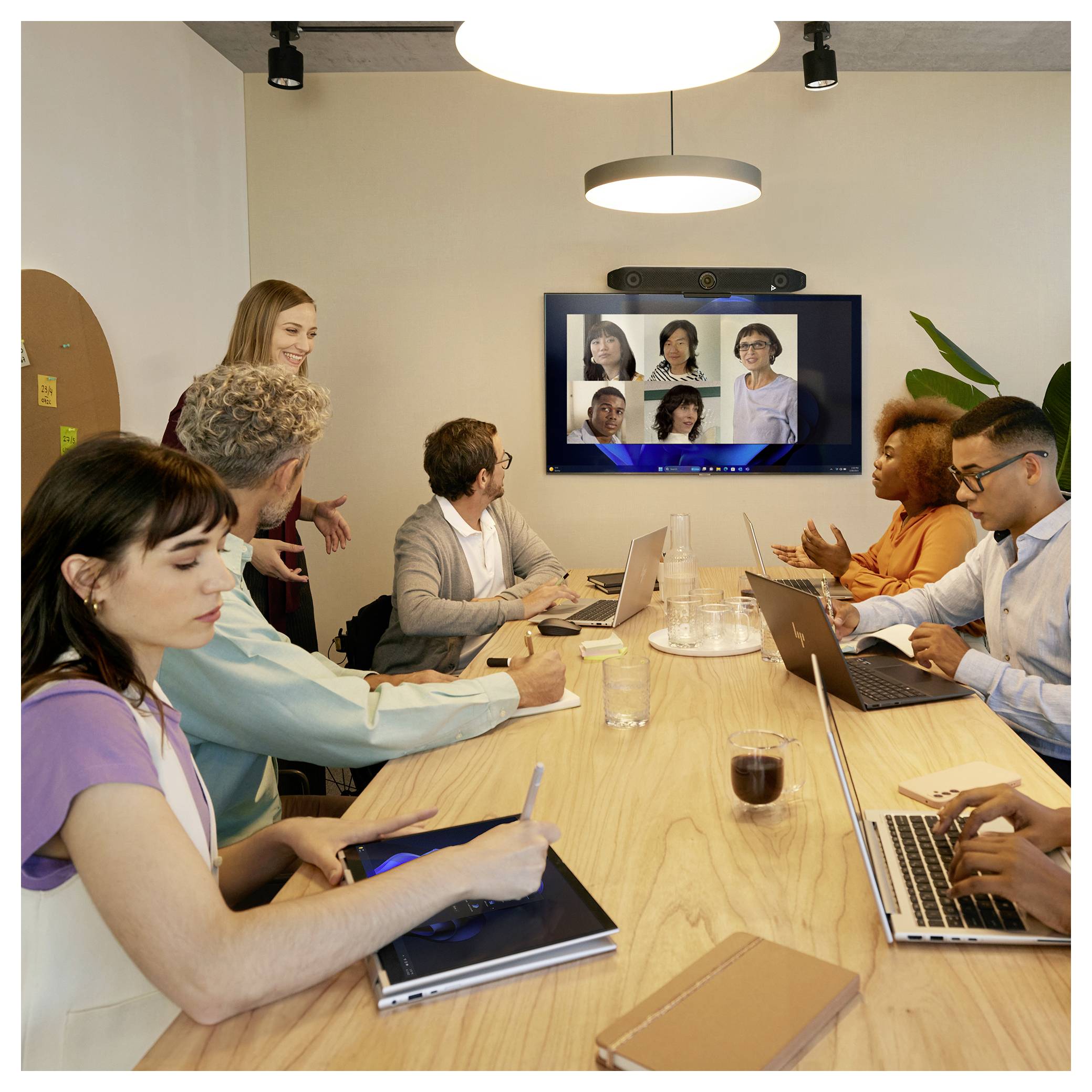Teammeeting rund um einen Konferenztisch mit mehreren Personen, die Laptops nutzen und einen Wandbildschirm beobachten, auf dem ein Remote-Videoanruf mit fünf Teilnehmern zu sehen ist.