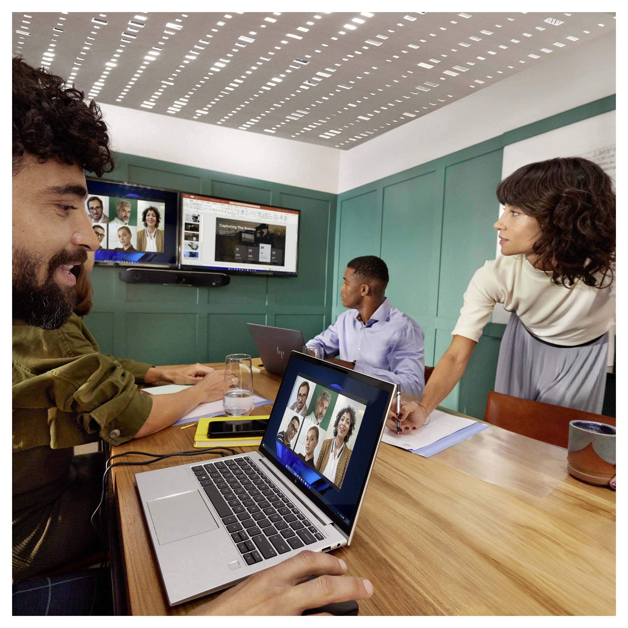 Teammeeting in einem Konferenzraum mit einem Videoanruf auf Laptops und Wandbildschirmen; vier Kollegen arbeiten zusammen an einem Tisch.