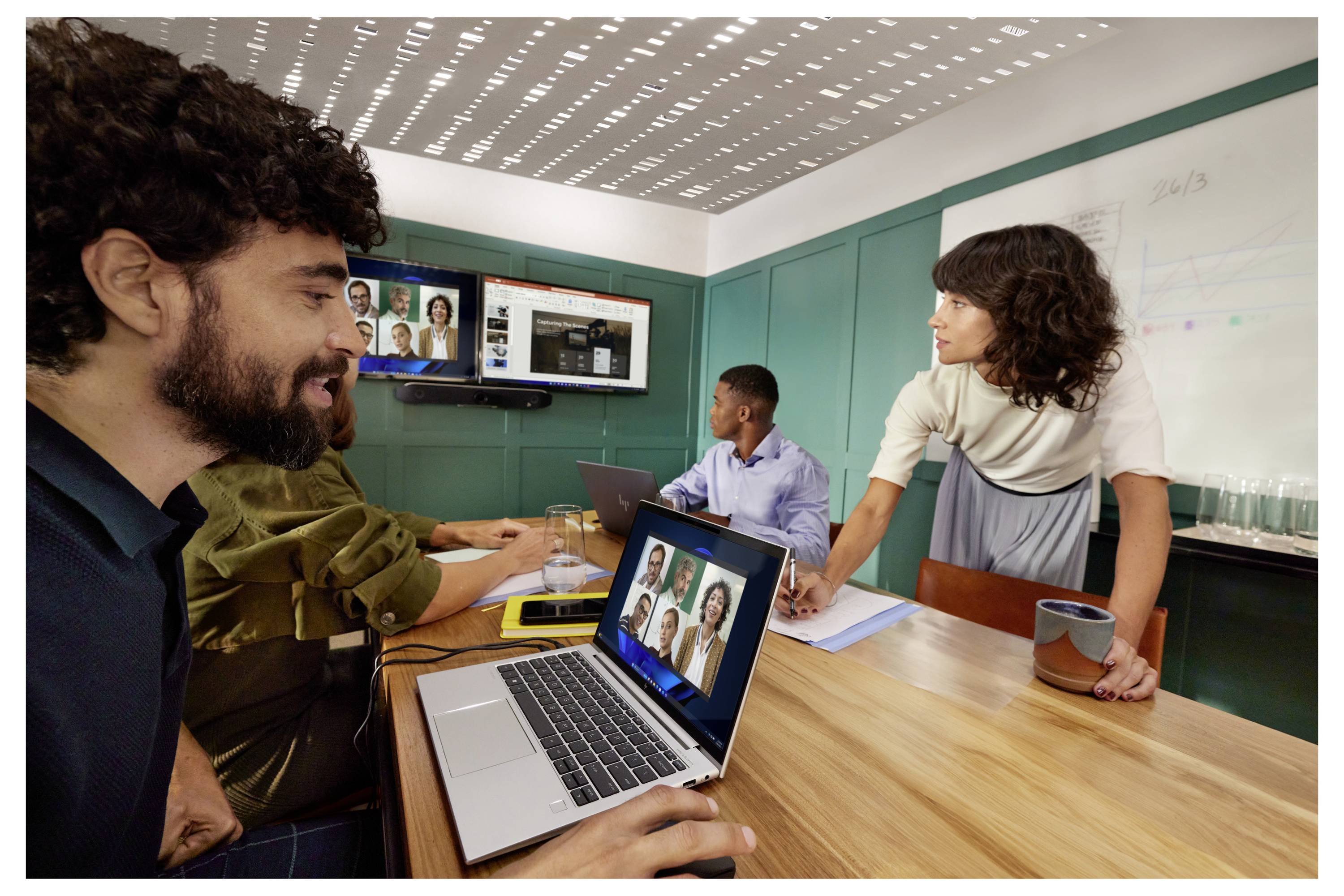 Menschen in einem Besprechungsraum arbeiten gemeinsam an einem Konferenztisch mit Laptops und einer Wandanzeige, die einen Videoanruf zeigt.