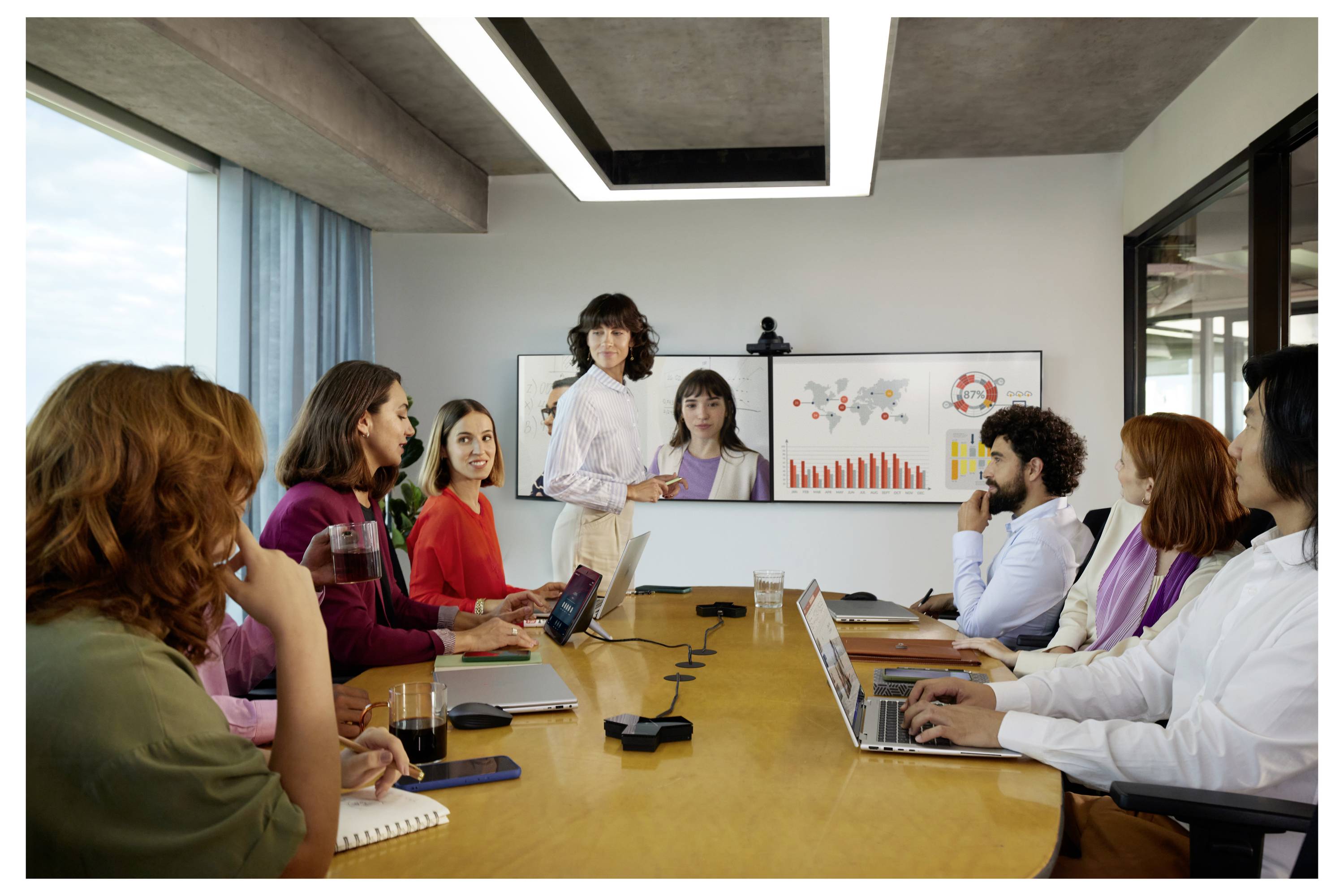 Geschäftstreffen mit einem Präsentator, der auf ein Whiteboard zeigt, das Diagramme anzeigt, während Kollegen um einen Konferenztisch herum mit Laptops und Notizbüchern sitzen.