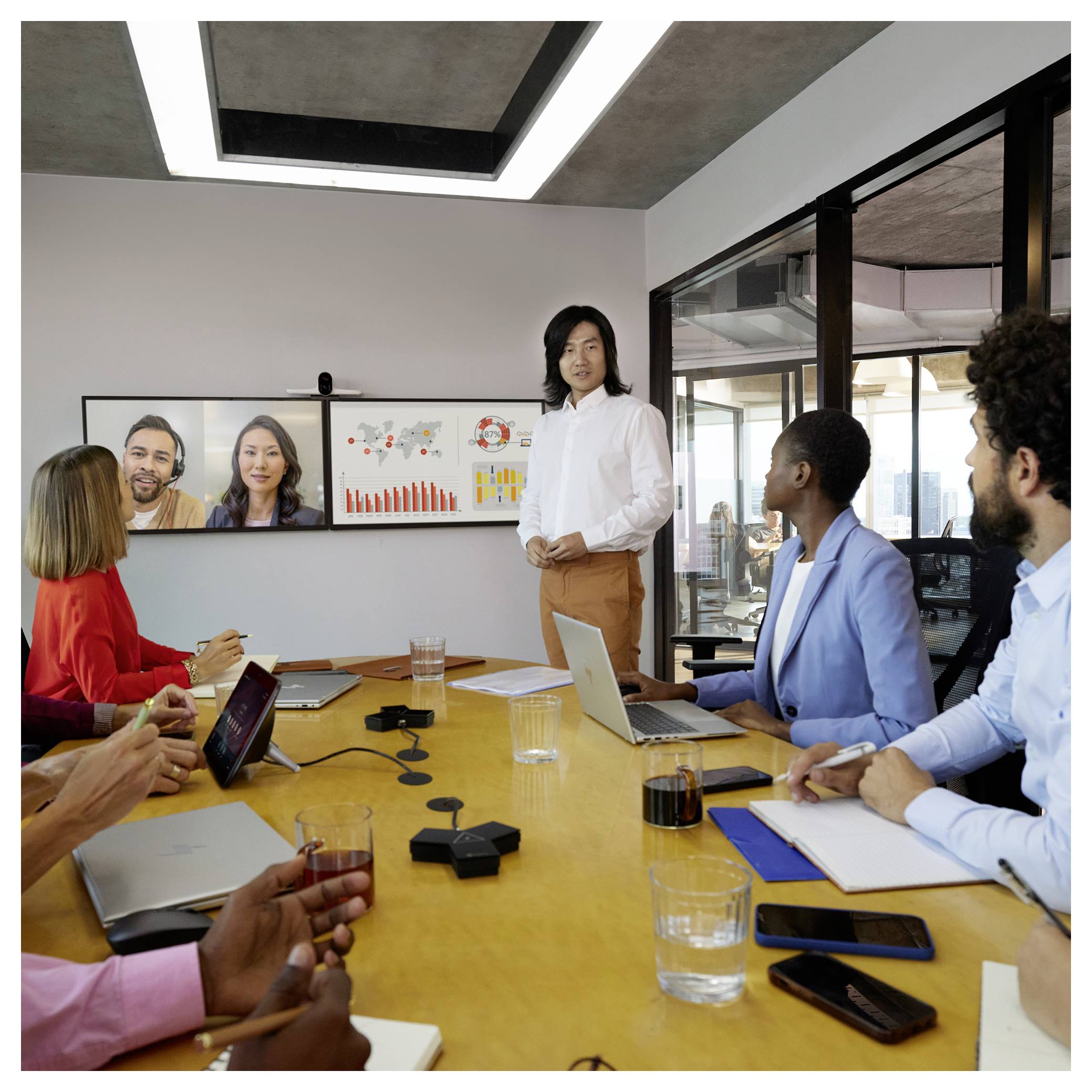 Geschäftstreffen mit einem Präsentator, der neben Wandbildschirmen mit Diagrammen steht, während Teammitglieder um einen Konferenztisch sitzen und zuschauen.