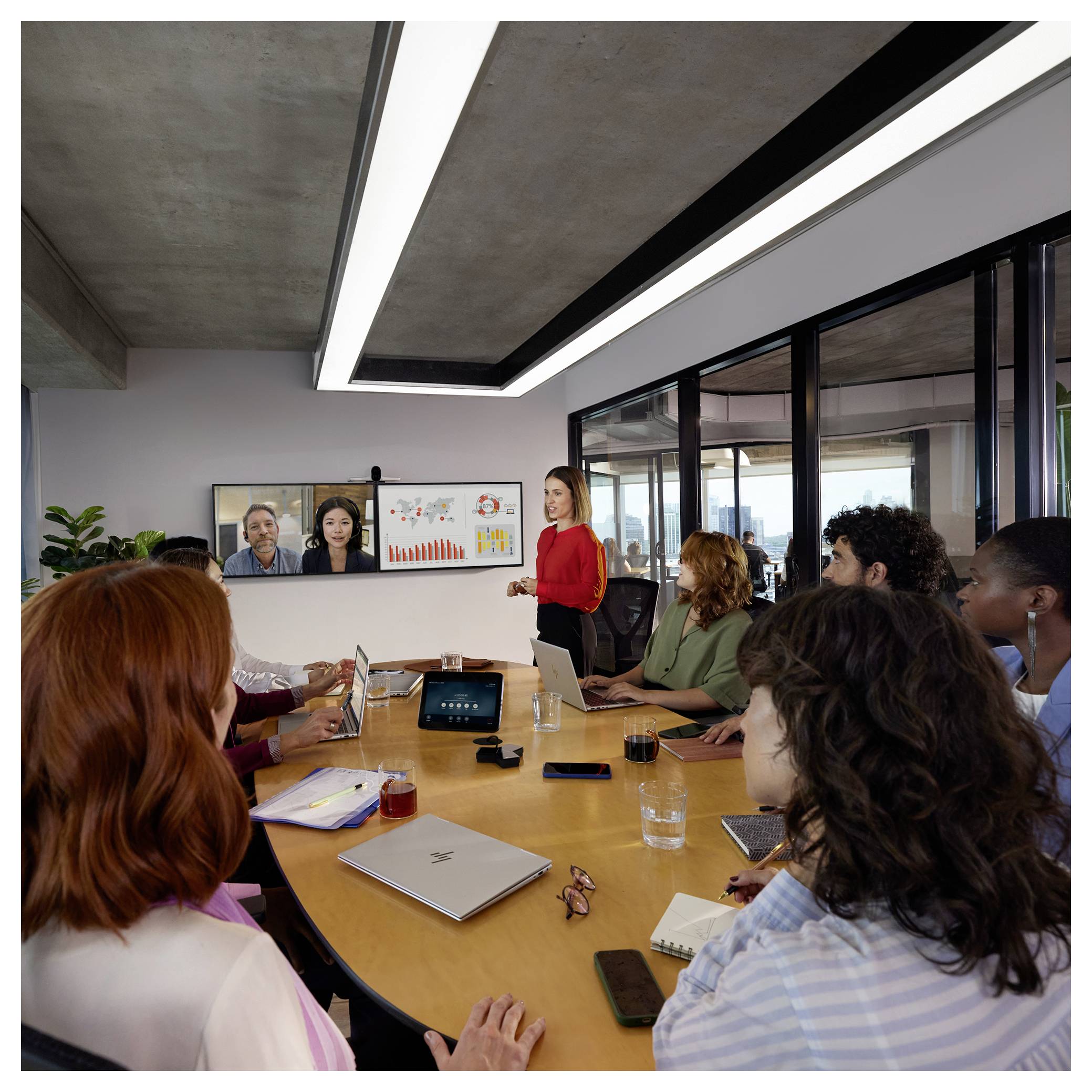 Geschäftstreffen mit einem Moderator in Rot, der mit Kollegen um einen Konferenztisch spricht, während zwei Remote-Teilnehmer auf einem Bildschirm zu sehen sind und Diagramme angezeigt werden.