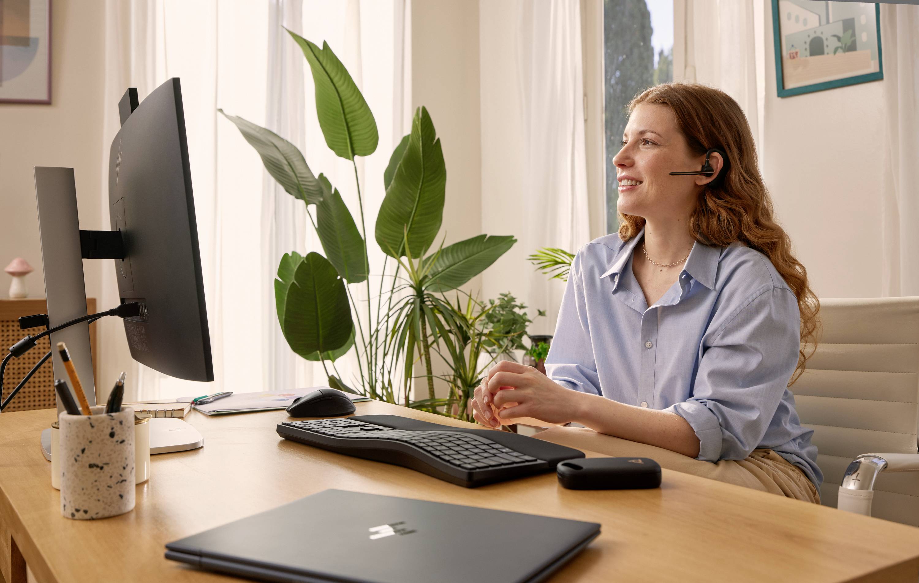 Frau mit Headset lächelt während eines Videoanrufs an einem Heimburotisch mit Computer, Tastatur, Laptop und Pflanzen.