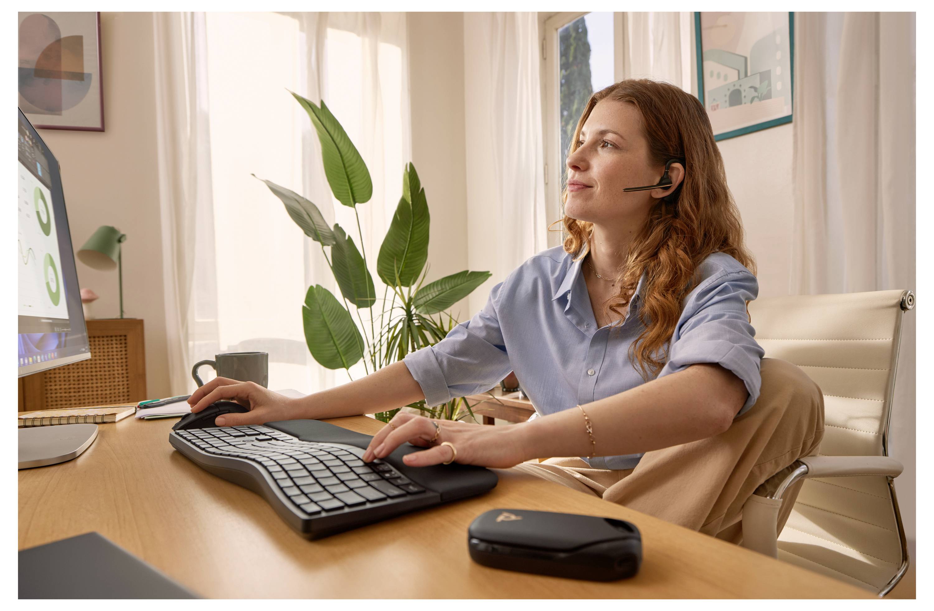 Frau mit Kopfhörer arbeitet an einem Schreibtisch an einem Computer in einem hellen Homeoffice und benutzt eine Tastatur und eine Maus.