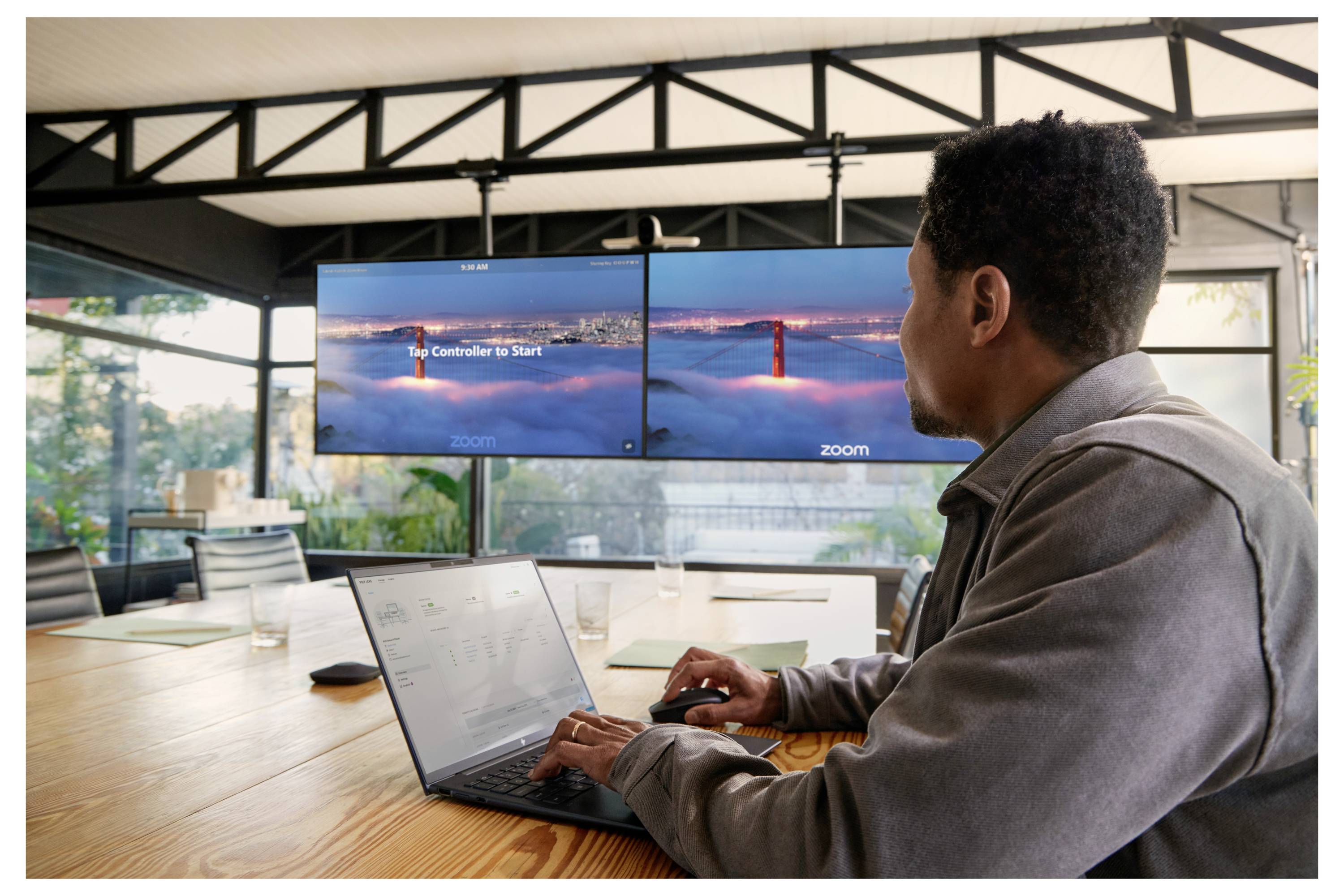 Ein Mann benutzt einen Laptop an einem Konferenztisch, während er auf zwei Monitoren ein Zoom-Meeting mit Blick auf die Golden Gate Bridge anschaut.