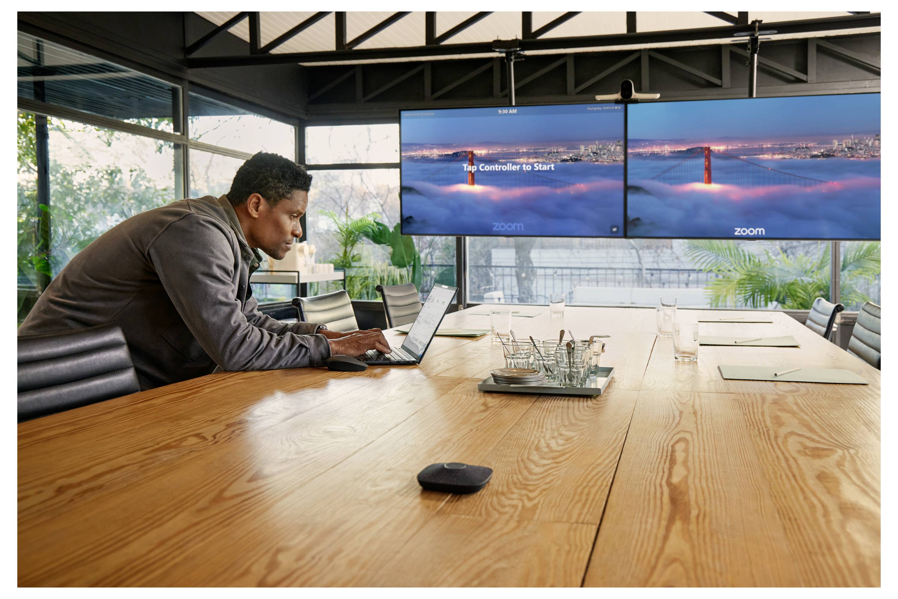 Ein Mann, der an einem Laptop an einem Konferenztisch in einem modernen Besprechungsraum arbeitet, mit zwei großen Zoom-Bildschirmen, auf denen 