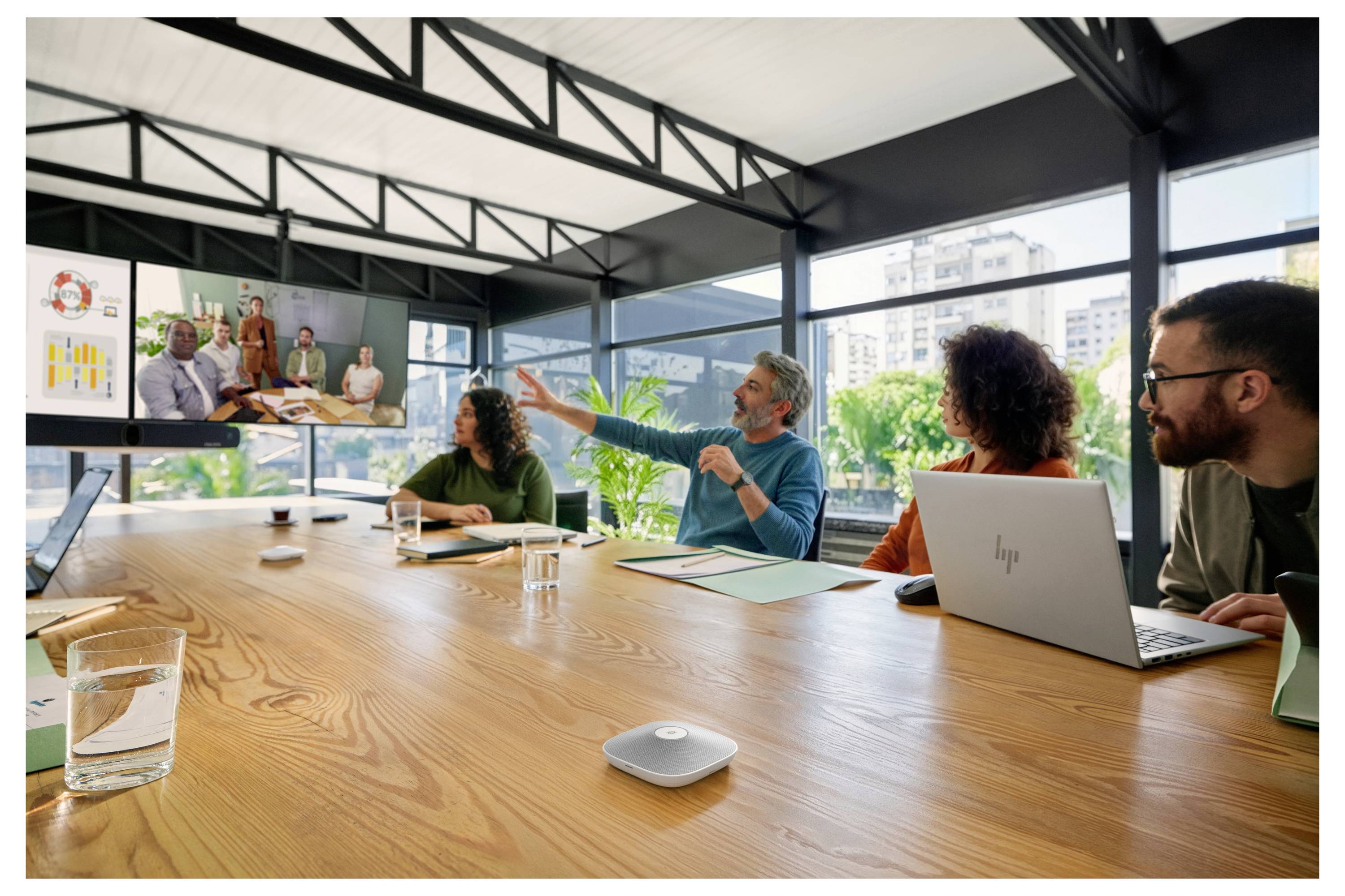 Teammeeting in einem modernen Konferenzraum, mit vier Personen um einen Tisch, die sich per Videokonferenz mit entfernten Kollegen auf einem Wandbildschirm abstimmen.