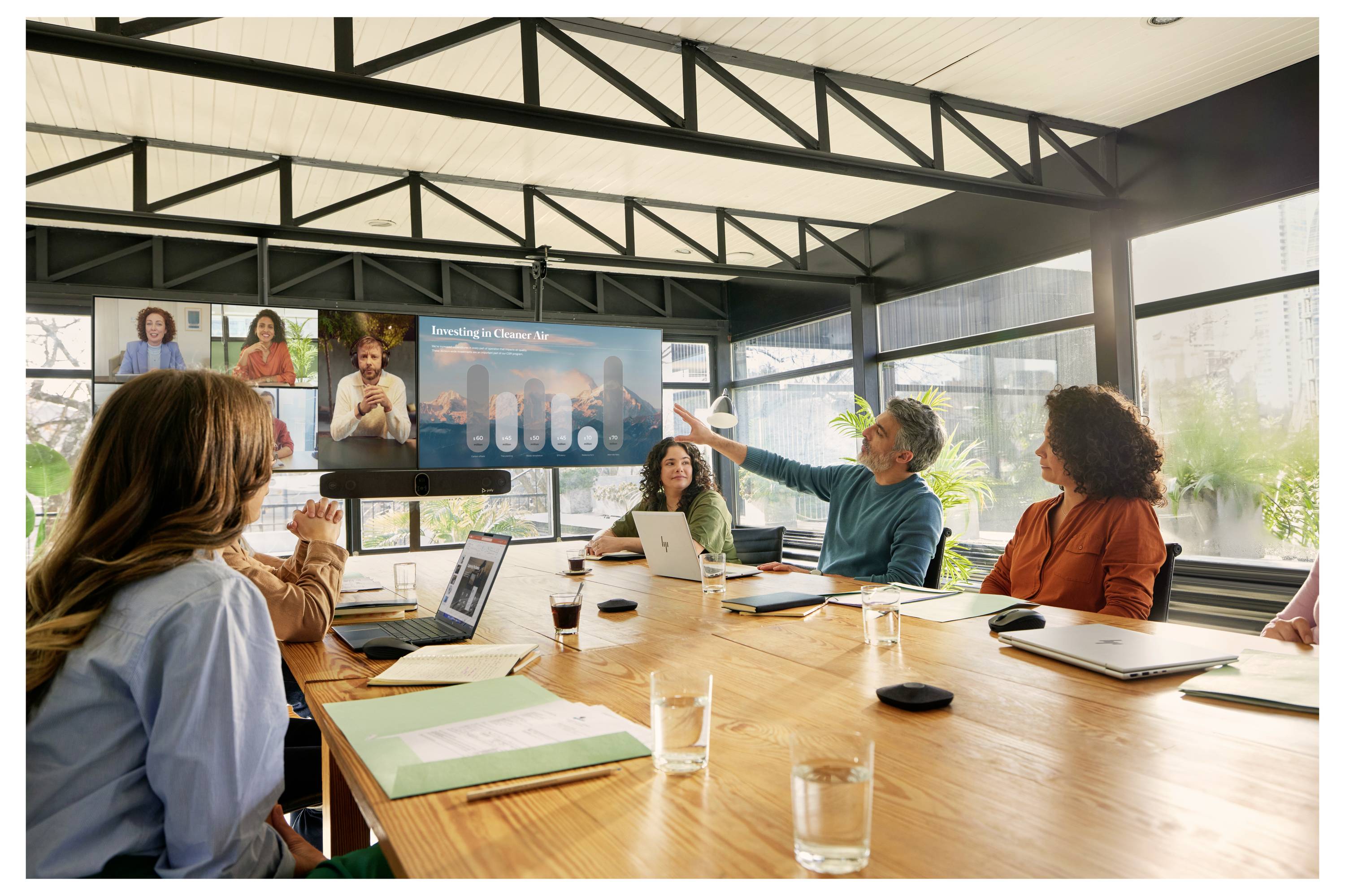 Teammeeting in einem Konferenzraum, bei dem ein Präsentator auf einen Bildschirm zeigt, der Fernbeteiligung und Diagramme anzeigt, während andere um einen Tisch herum zuhören.