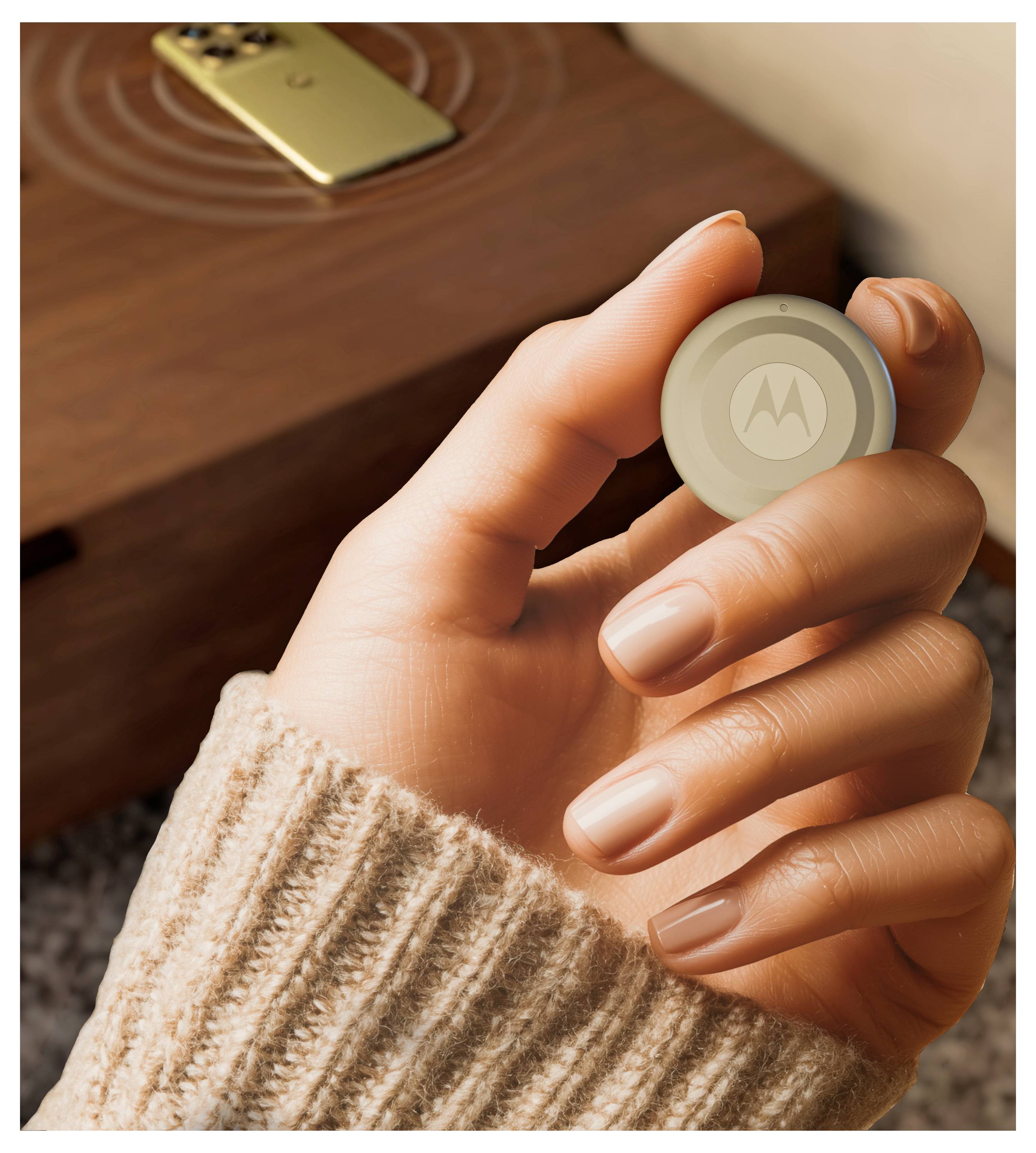Eine Hand hält ein kleines, rundes Motorola-Gerät. Im Hintergrund liegt ein Smartphone auf einer Holzoberfläche, was auf ein Technologie-Setup hindeutet.