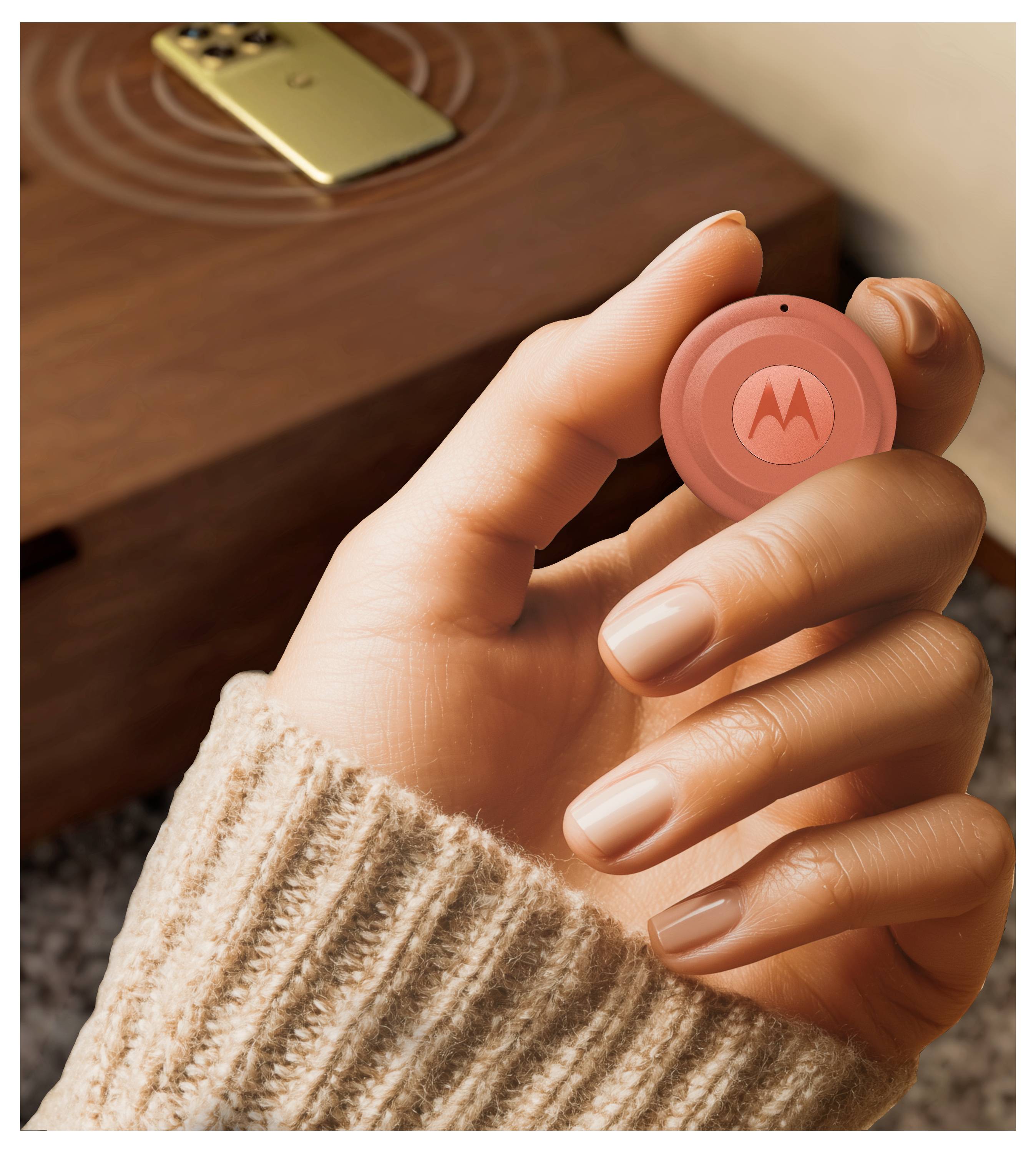 Eine Hand hält ein rundes, orangefarbenes elektronisches Gerät mit dem Motorola-Logo, was auf die Verwendung als drahtloser Tracker hindeutet. Ein Smartphone ruht auf einer Holzoberfläche im Hintergrund.