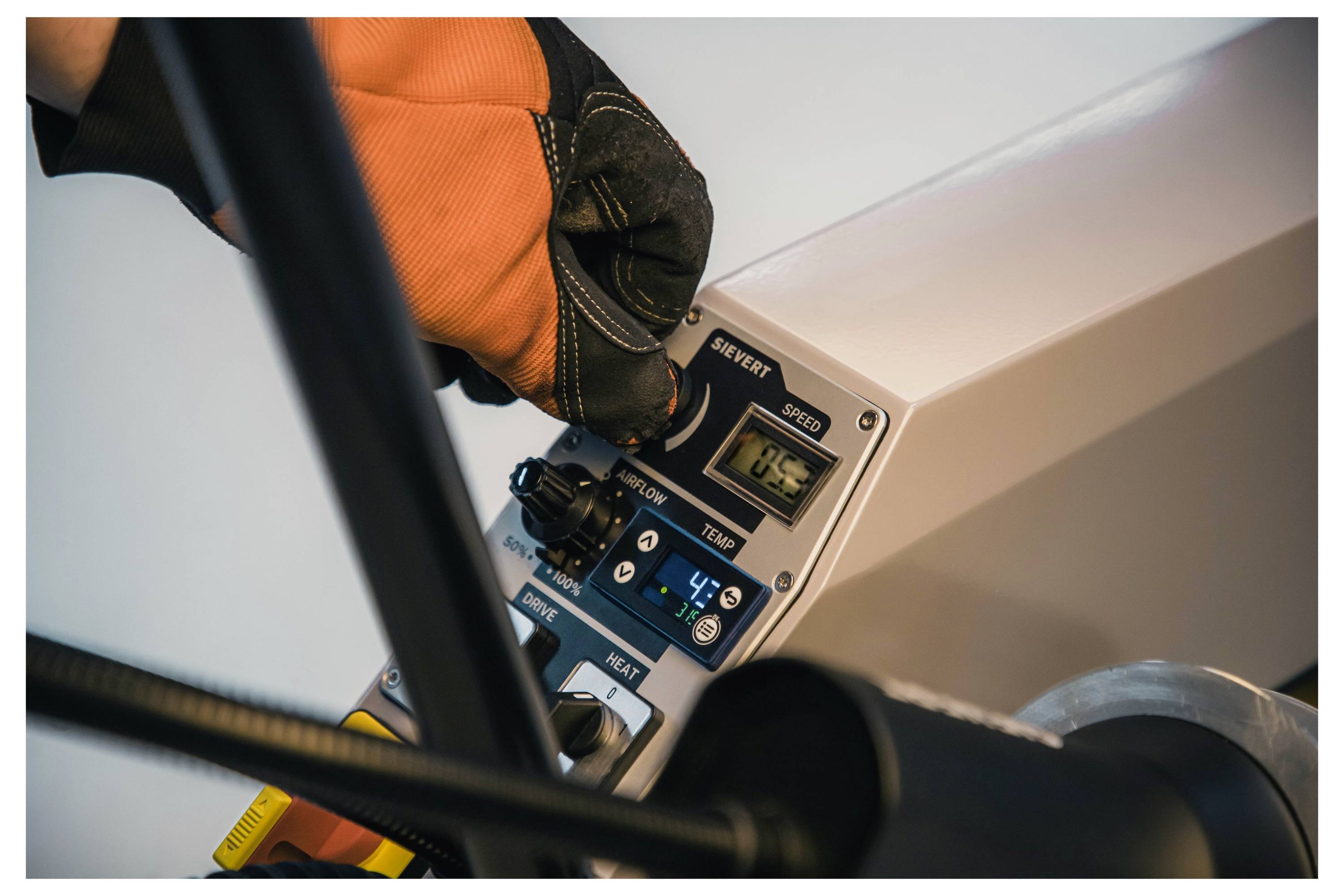 Nahaufnahme einer Person, die einen orangefarbenen Handschuh trägt und ein Bedienfeld an einer Industriemaschine mit Einstellungen und Zifferblättern anpasst.