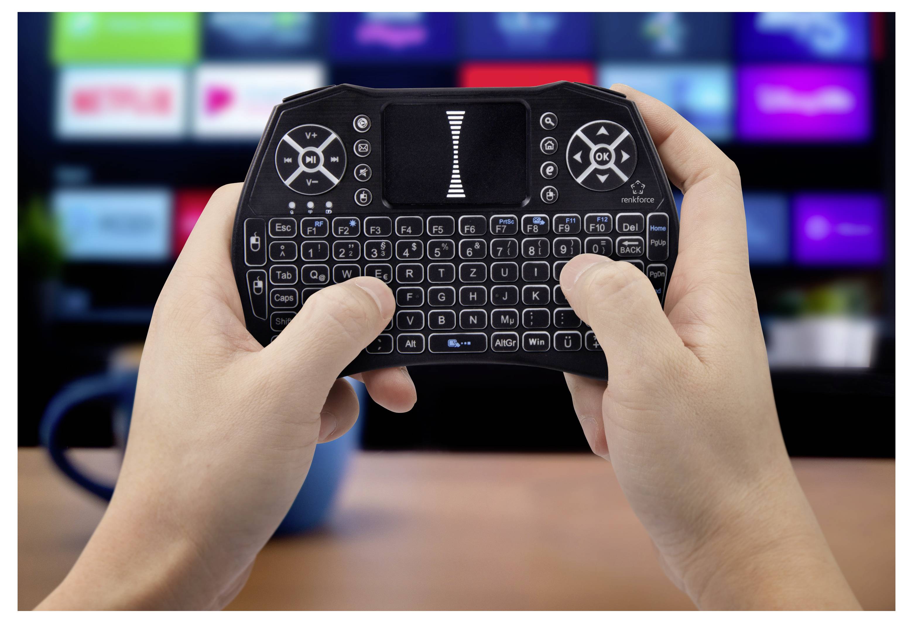 Hände, die eine Mini-Wireless-Tastatur mit Touchpad halten, vor einem unscharfen Fernseh­bildschirm, was auf die Fernbedienung von Medien oder die Navigation auf Smart-TVs hindeutet.