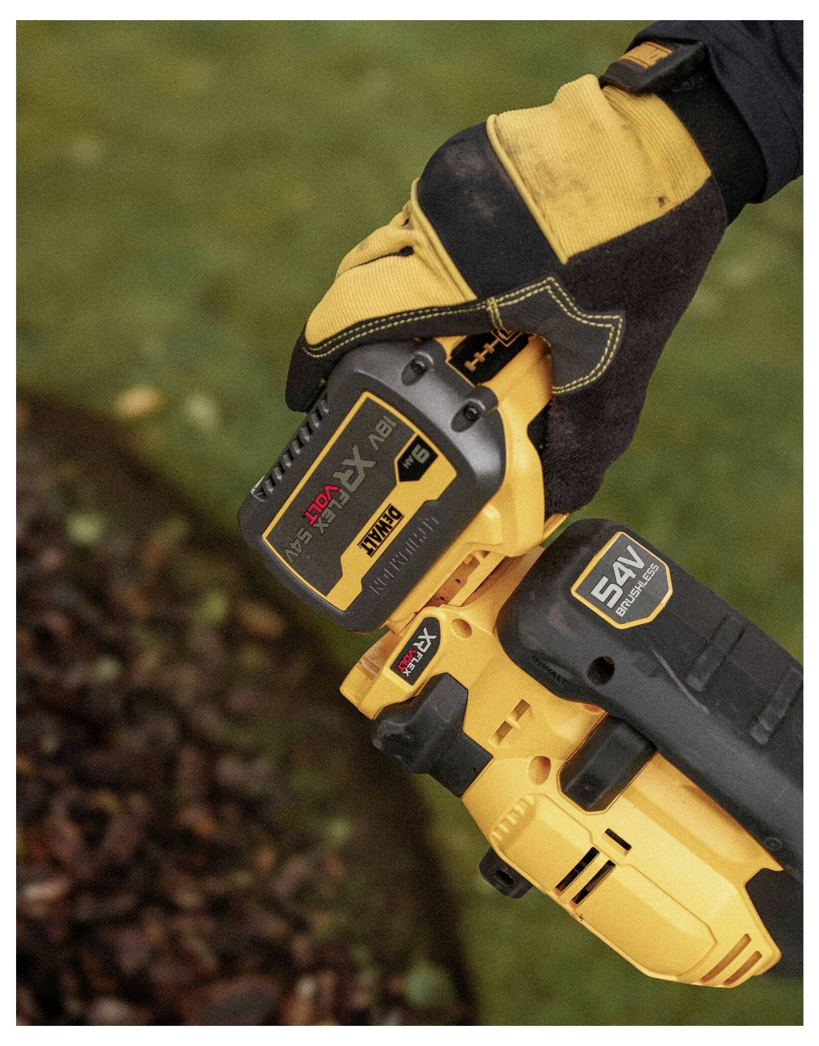 Eine Person, die einen Handschuh trägt, bedient einen gelben kabellosen Laubblaser und hält dabei die 54-V-Batterie. Der Boden ist mit Blättern bedeckt.
