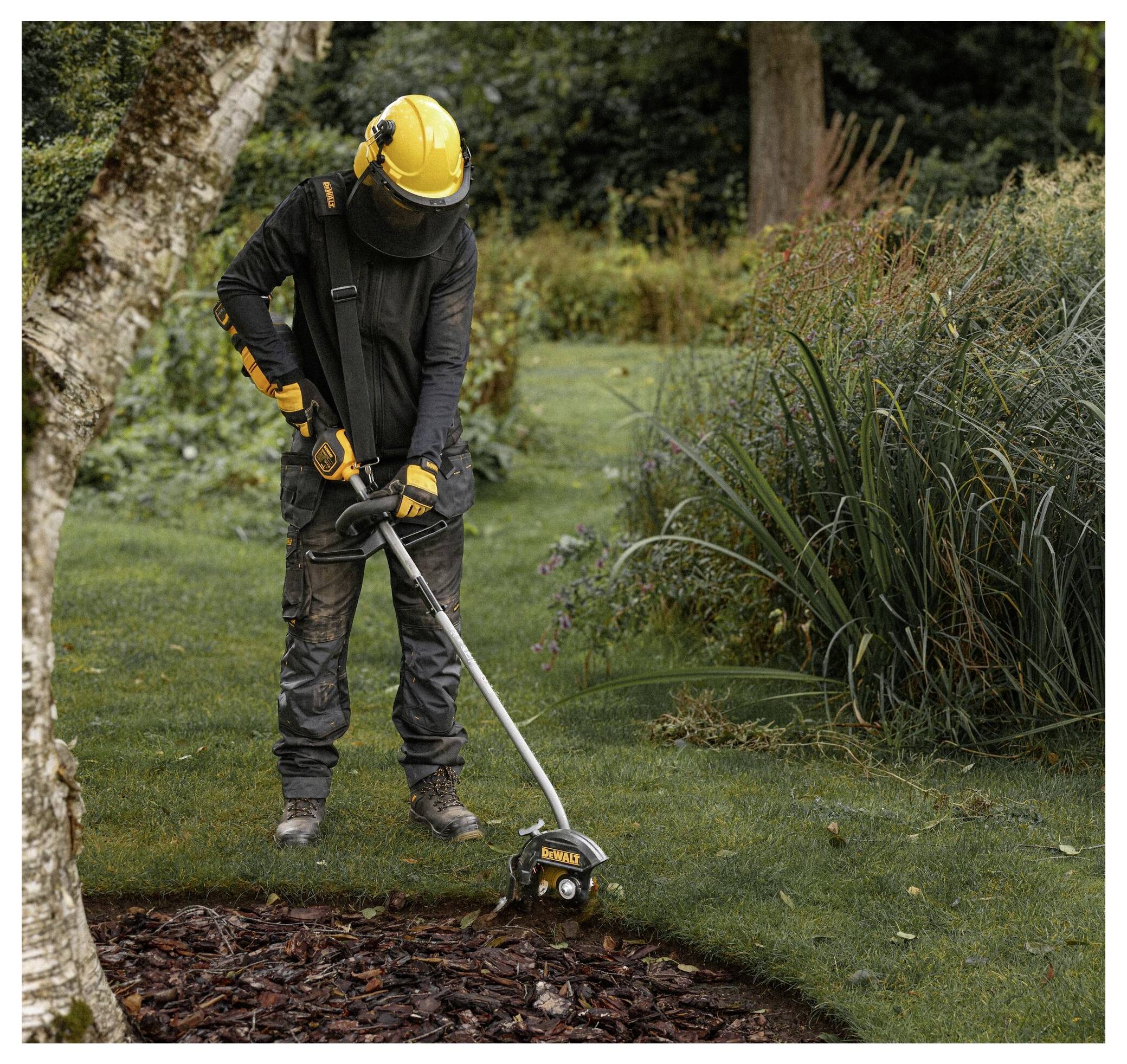 Eine Person in Schutzausrüstung trimmt mit einem Elektrowerkzeug Gras in der Nähe eines Gartenbeets, das von Rindenmulch umgeben ist, in einem angelegten Garten.