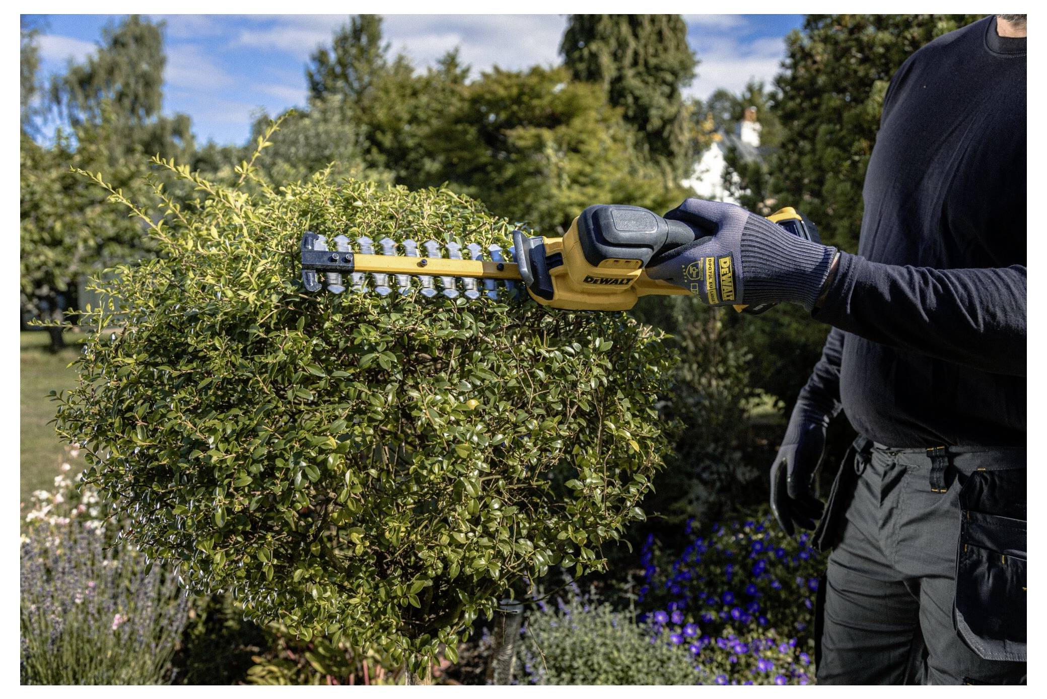 Eine Person schneidet einen Strauch mit einer gelb-schwarzen elektrischen Heckenschere in einem Garten zurecht, umgeben von Bäumen und Blumen unter einem blauen Himmel.