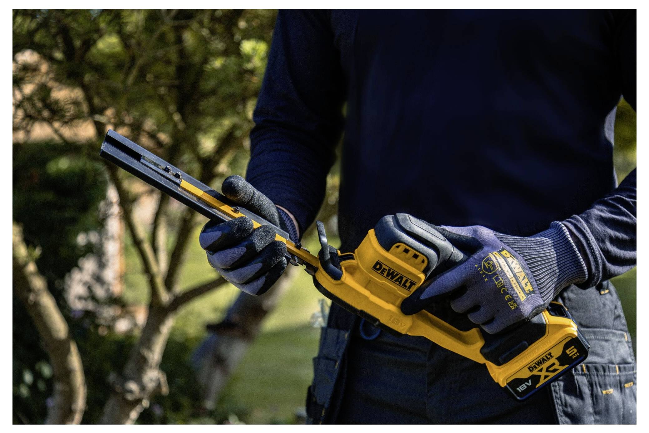 Eine Person mit Handschuhen bedient eine DeWalt-Reciprosäge im Freien, umgeben von Grünflächen, was die Werkzeugbedienung demonstriert.