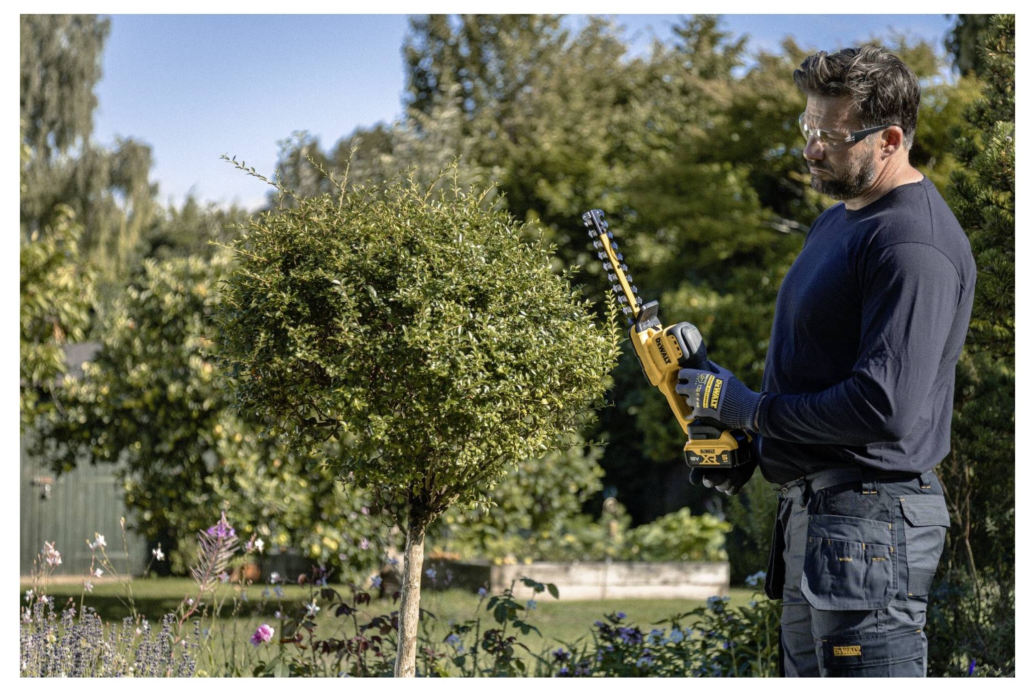 Eine Person in Schutzbrille schneidet Formgehölze mit einer kabellosen Heckenschere in einem Garten, mit Bäumen und Sträuchern im Hintergrund.