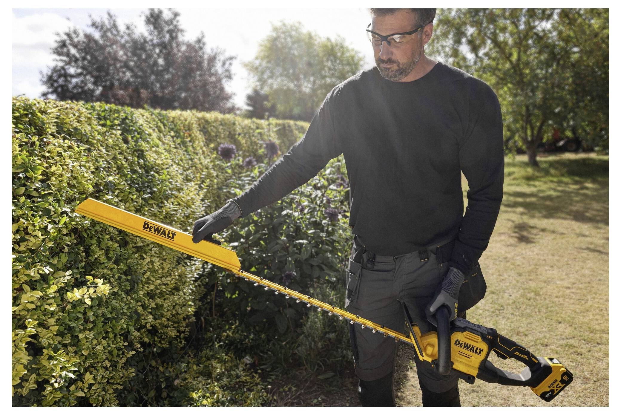 Eine Person in Schutzausrüstung verwendet einen gelben Heckenschneider, um eine Gartenhecke unter sonnigem Himmel zu formen.