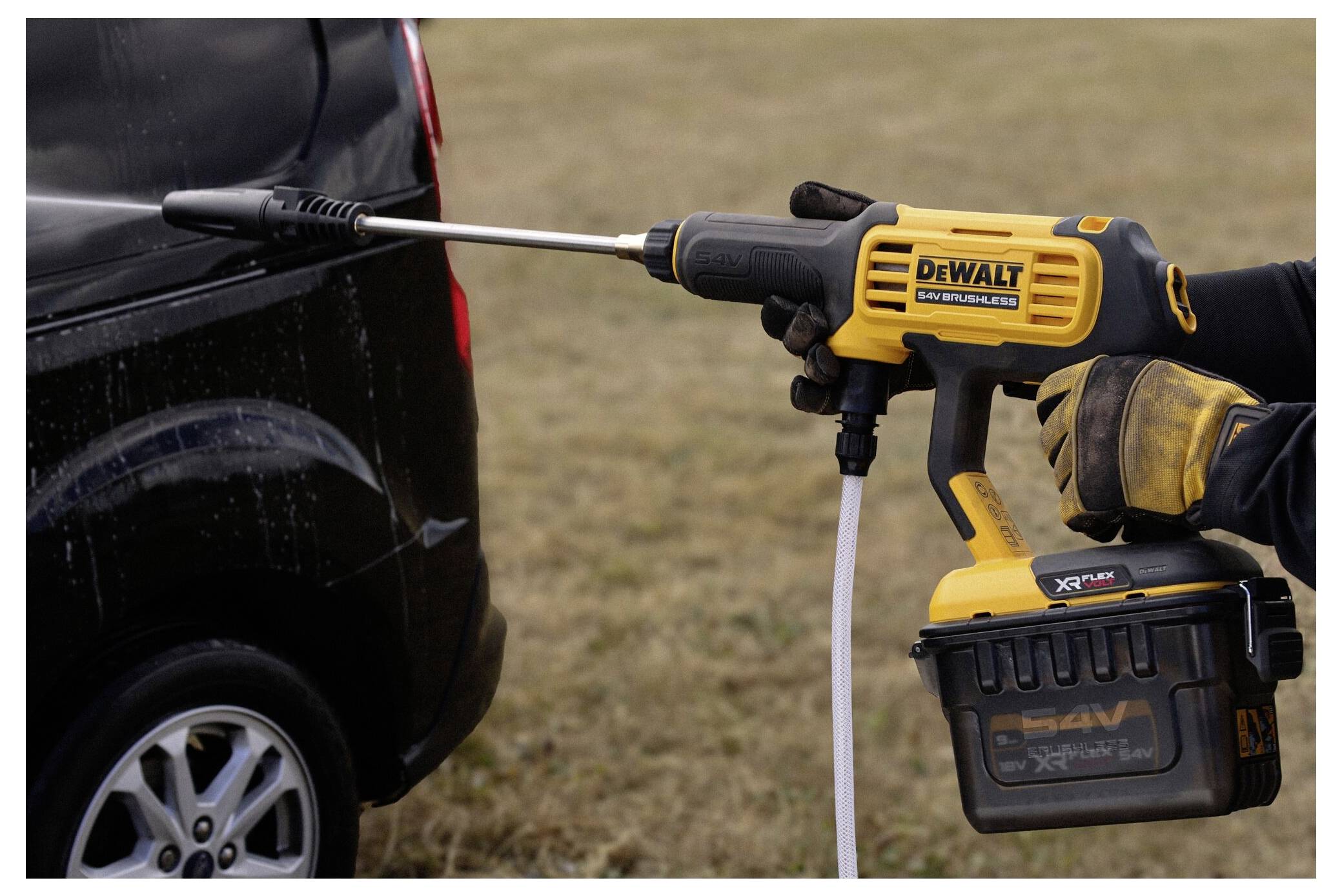 Eine Person benutzt einen gelben kabellosen Hochdruckreiniger, um ein schwarzes Fahrzeug in einer Außenumgebung zu reinigen.