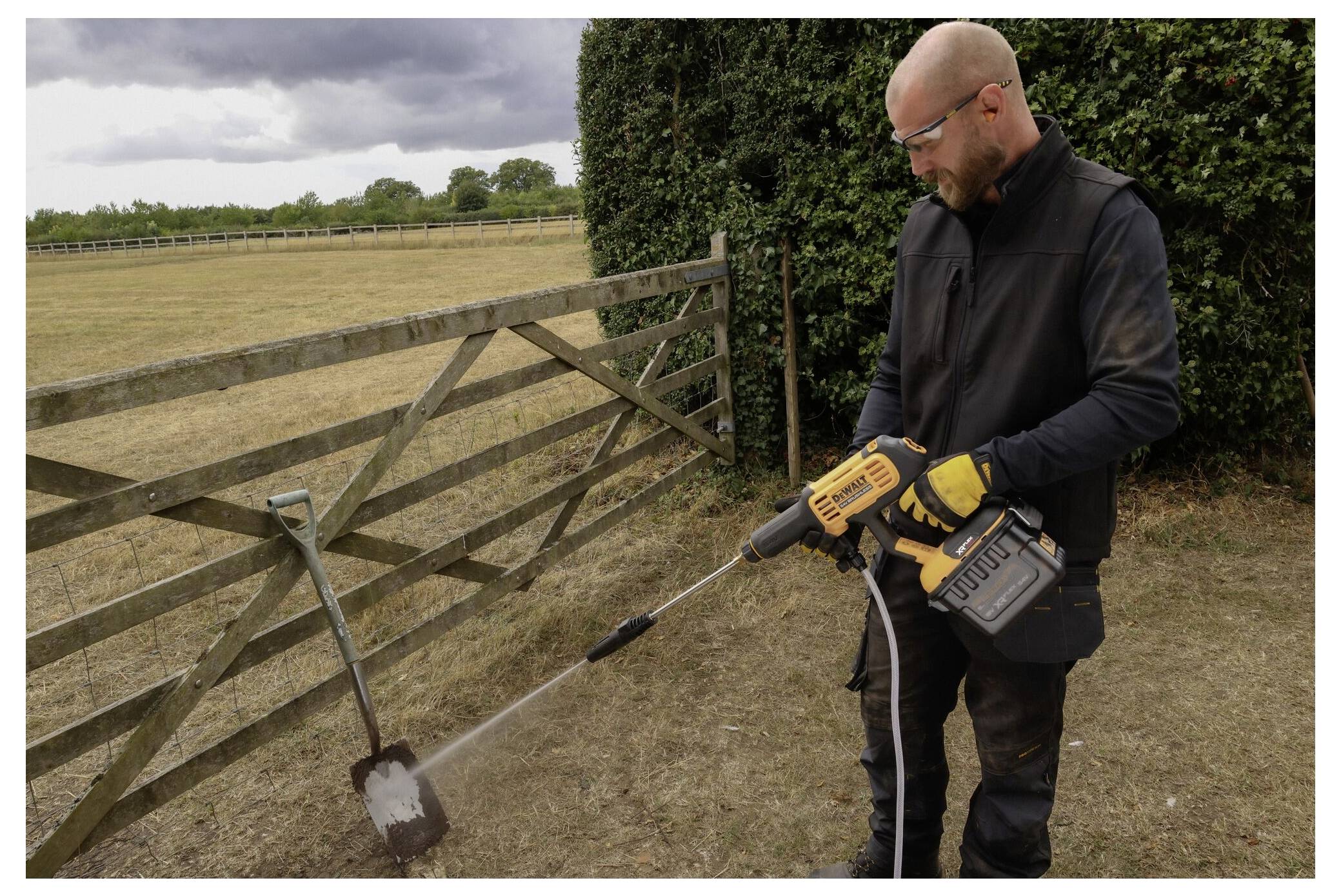 Eine Person in Schutzbrille benutzt einen Hochdruckreiniger, um eine Schaufel im Freien zu reinigen, neben einem Holzzaun und einer Hecke, an einem bewölkten Tag.