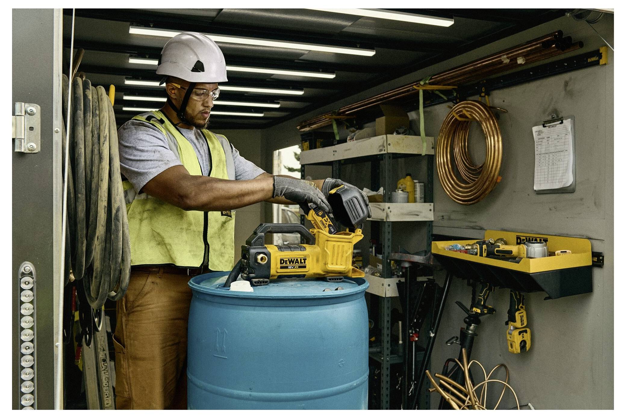 Ein Arbeiter, der Schutzausrüstung und einen Helm trägt, bedient ein Elektrowerkzeug in einer Werkstatt mit Regalen voller Ausrüstung und einer an der Wand befestigten Zwischenablage.