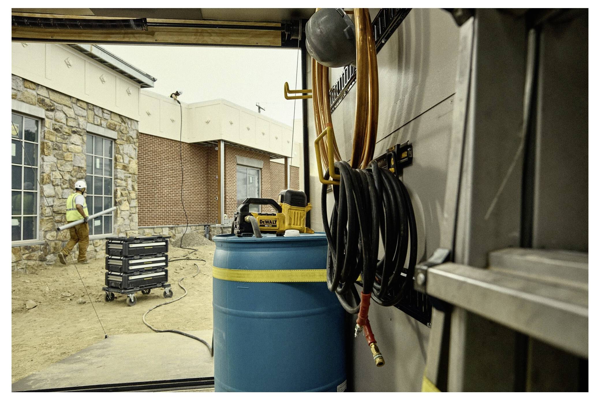 Ein Bauarbeiter sprüht Wasser auf die Erde neben einem Backsteingebäude. Die Innenansicht zeigt einen Schlauch, der an der Wand eines Lastwagens aufgerollt ist.