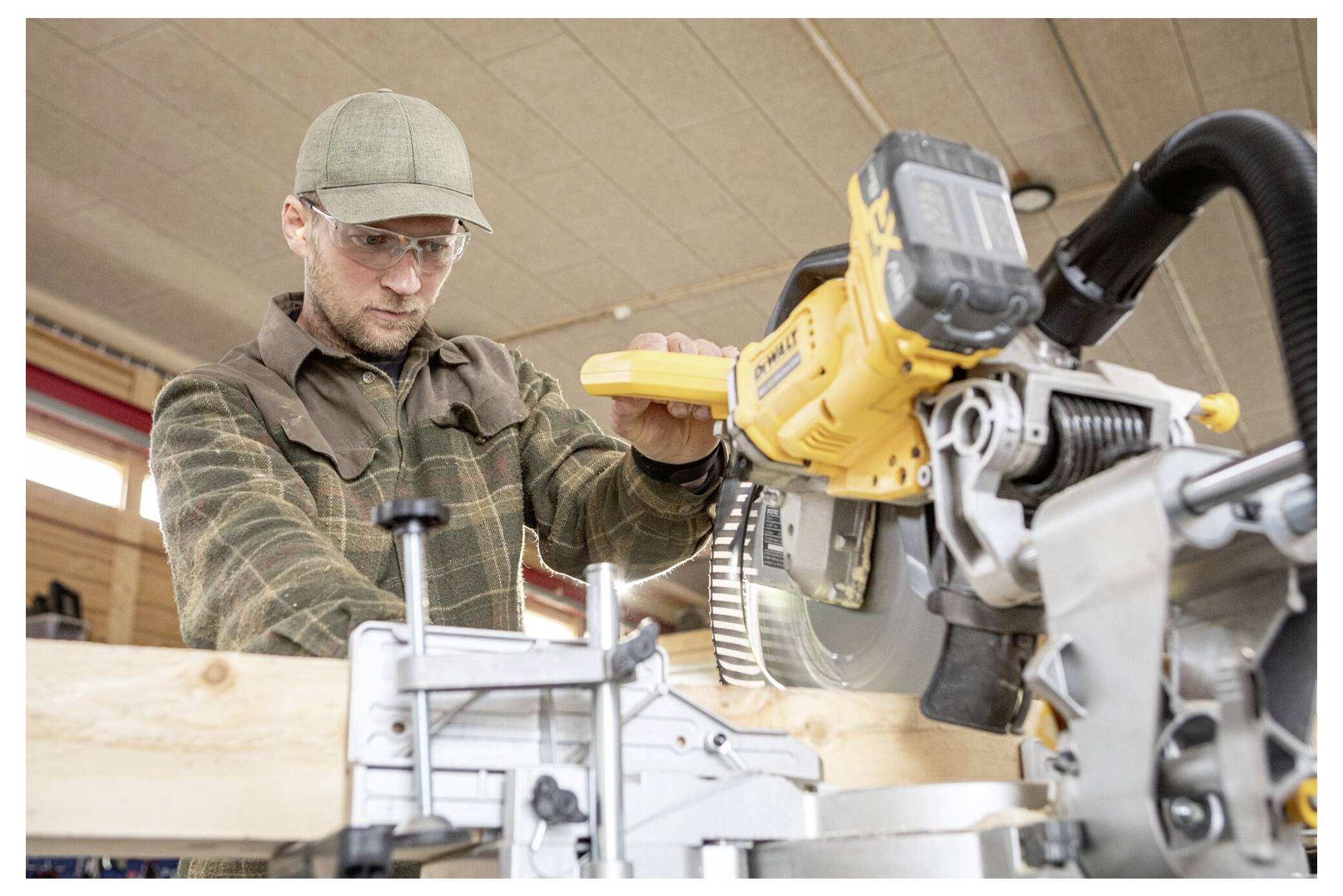 Eine Person mit Schutzbrille und Mütze bedient eine Kreissäge und schneidet ein Holzbrett in einer Werkstattumgebung.