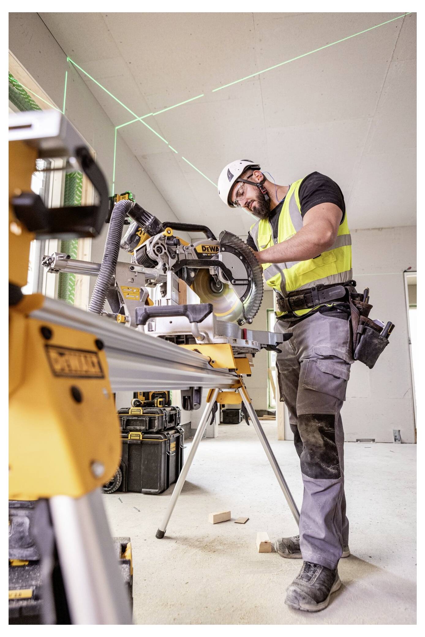 Ein Bauarbeiter in Schutzausrüstung bedient eine Gehrungssäge in einem teilweise fertiggestellten Raum mit bloßgelegten Wänden und verschiedenen Werkzeugen rundherum.