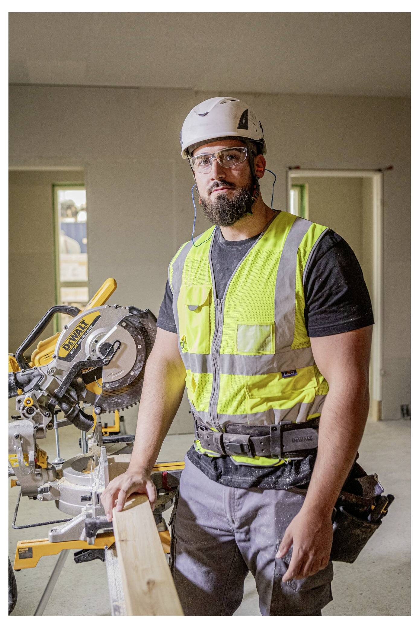 Ein Bauarbeiter in einer Sicherheitsweste und einem Schutzhelm steht neben einer Elektrosäge und hält ein Holzbrett in einem teilweise errichteten Raum.