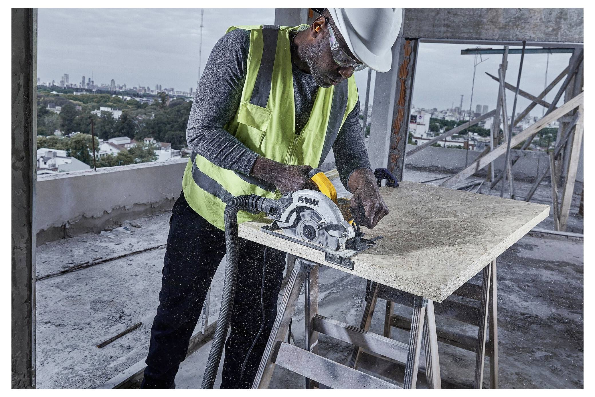 Ein Bauarbeiter in Helm und Warnweste benutzt eine Kreissäge, um Sperrholz auf einer Hochhausbaustelle zu schneiden, die eine Stadtlandschaft überblickt.