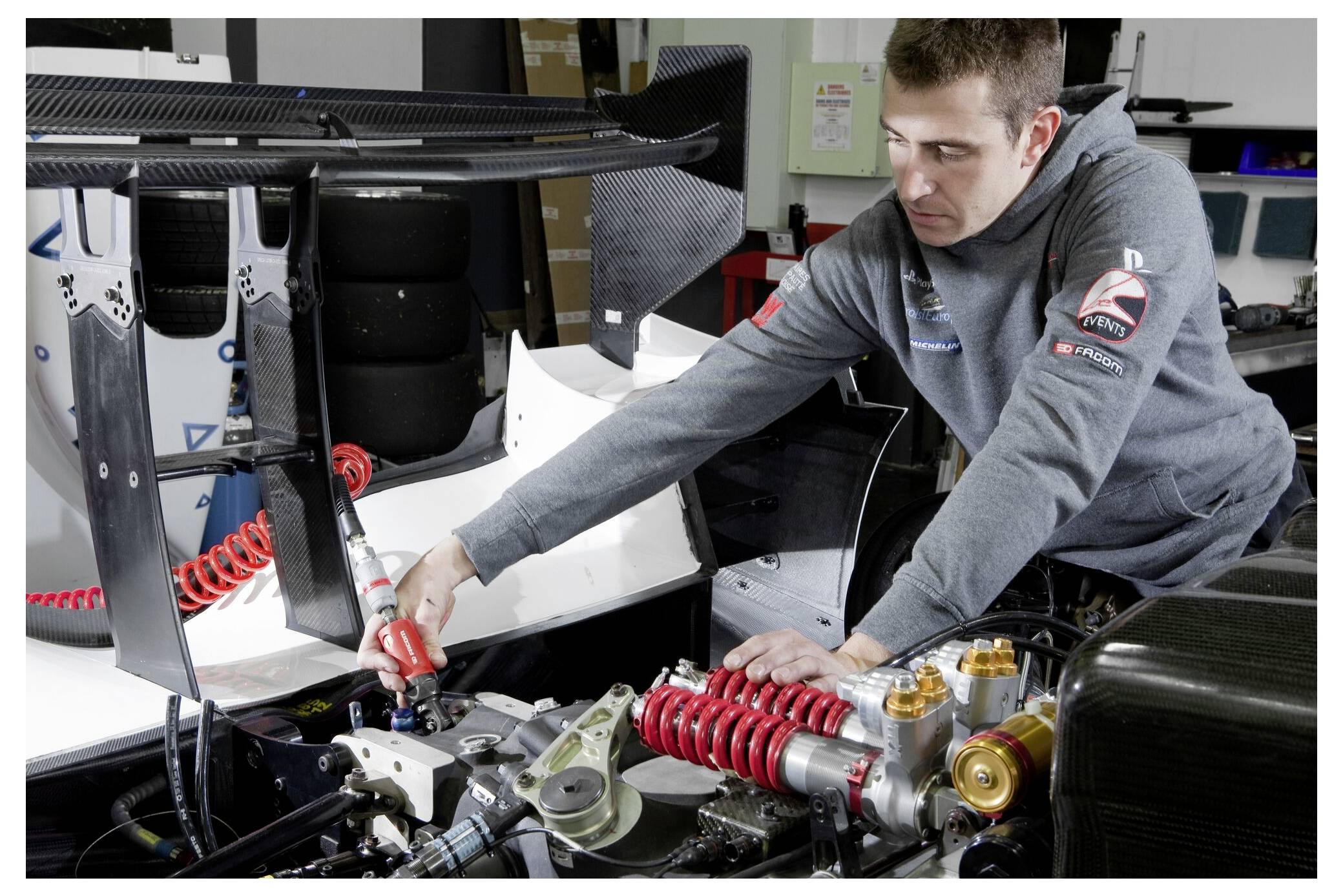 Ein Mechaniker arbeitet an einem Rennwagen und stellt Komponenten in der Nähe des Heckflügels und der Aufhängung ein, umgeben von Werkzeugen und Ausrüstung in einer Werkstatt.