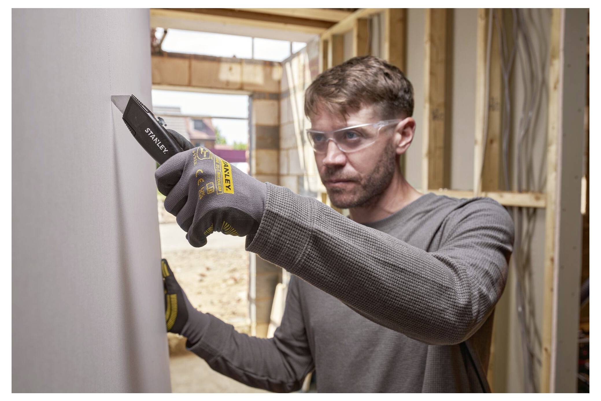 Eine Person, die Schutzbrille und Handschuhe trägt, verwendet ein Universalmesser, um Material in einem Baubereich mit freigelegtem Holzrahmen zu schneiden.