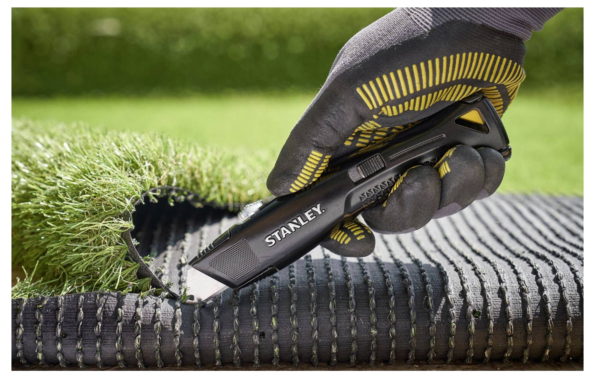 Eine Person mit behandschuhten Händen schneidet künstliches Gras mit einem Stanley-Universalmesser und zeigt einen präzisen, sauberen Schnitt in einer Gartenumgebung.