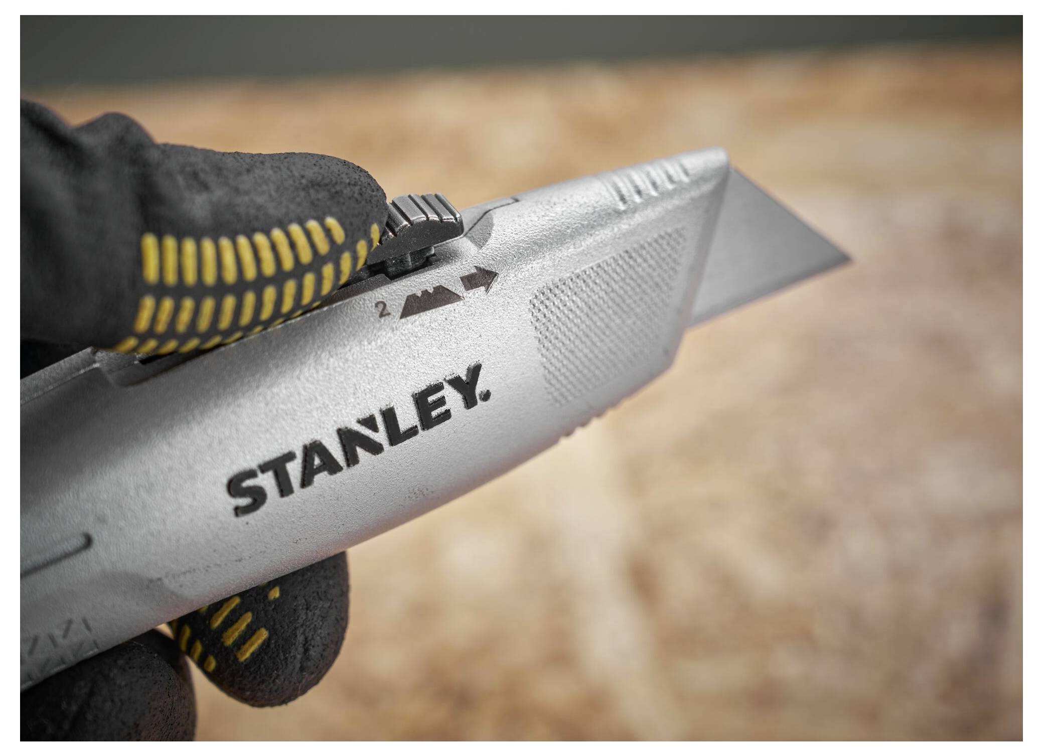 Eine behandschuhte Hand stellt die Klingenlänge eines Stanley-Universalmessers ein, in Nahaufnahme vor einem Holzoberflächenhintergrund dargestellt.