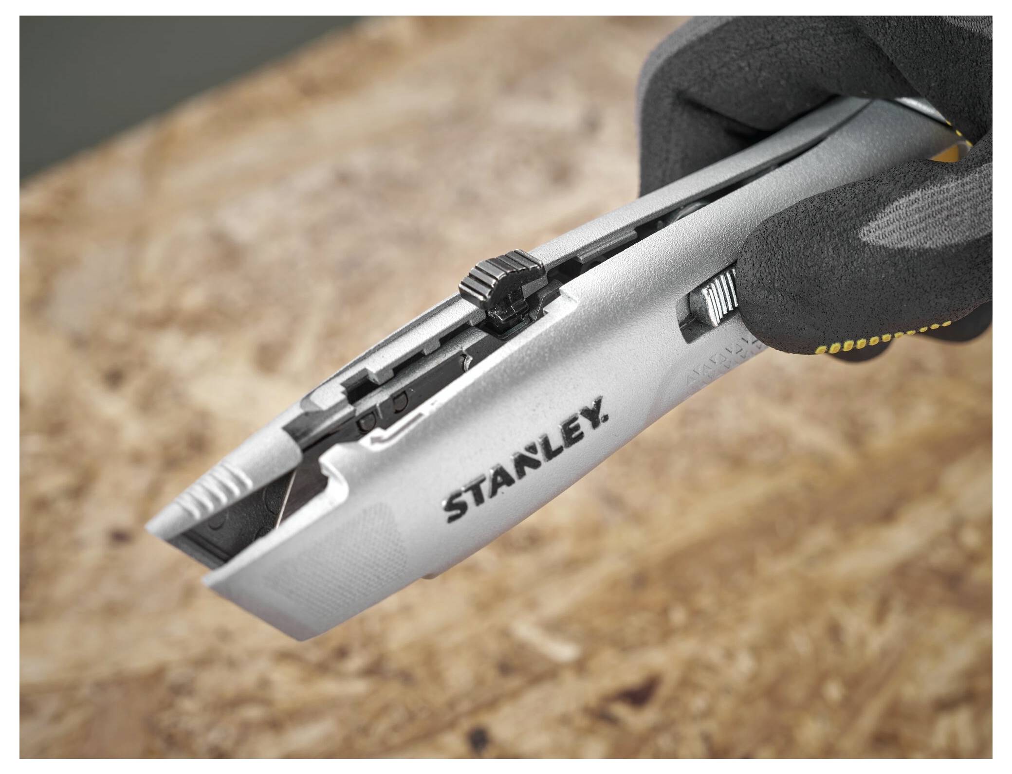 Eine behandschuhte Hand einer Person hält ein graues Universalmesser mit einziehbarer Klinge über einer Holzoberfläche.