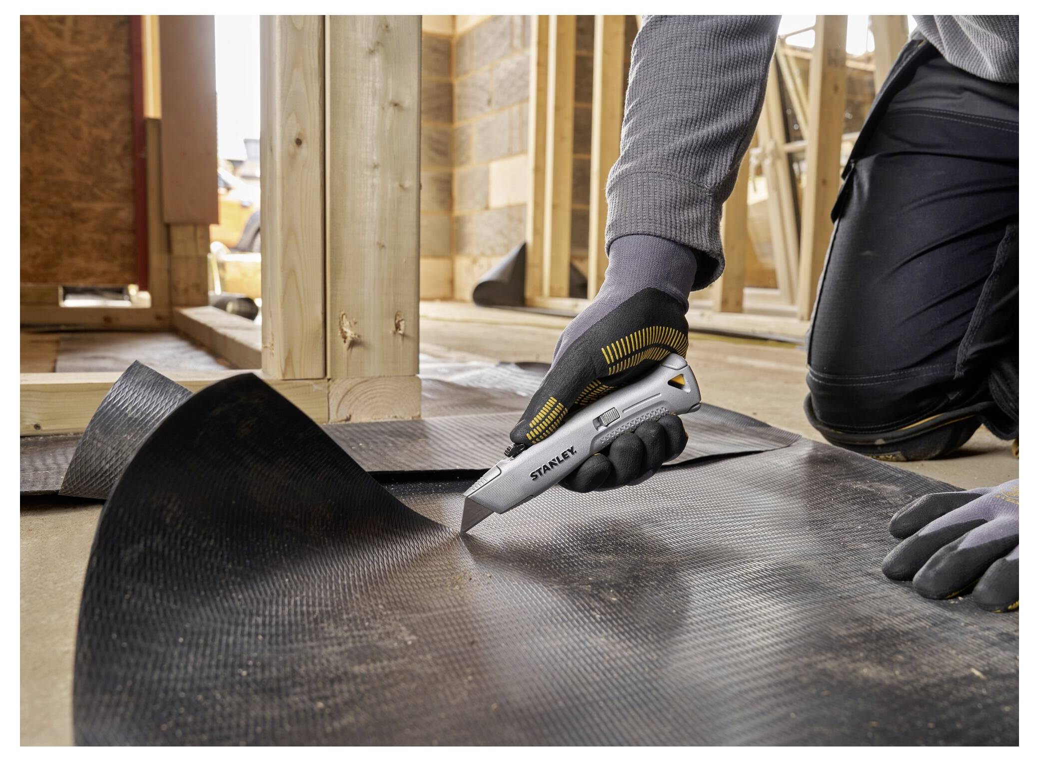 Eine Person in Arbeitskleidung verwendet ein Universalmesser, um schwarzes Bodenmaterial in einer Baustelle zu schneiden, mit Holzbalken im Hintergrund.