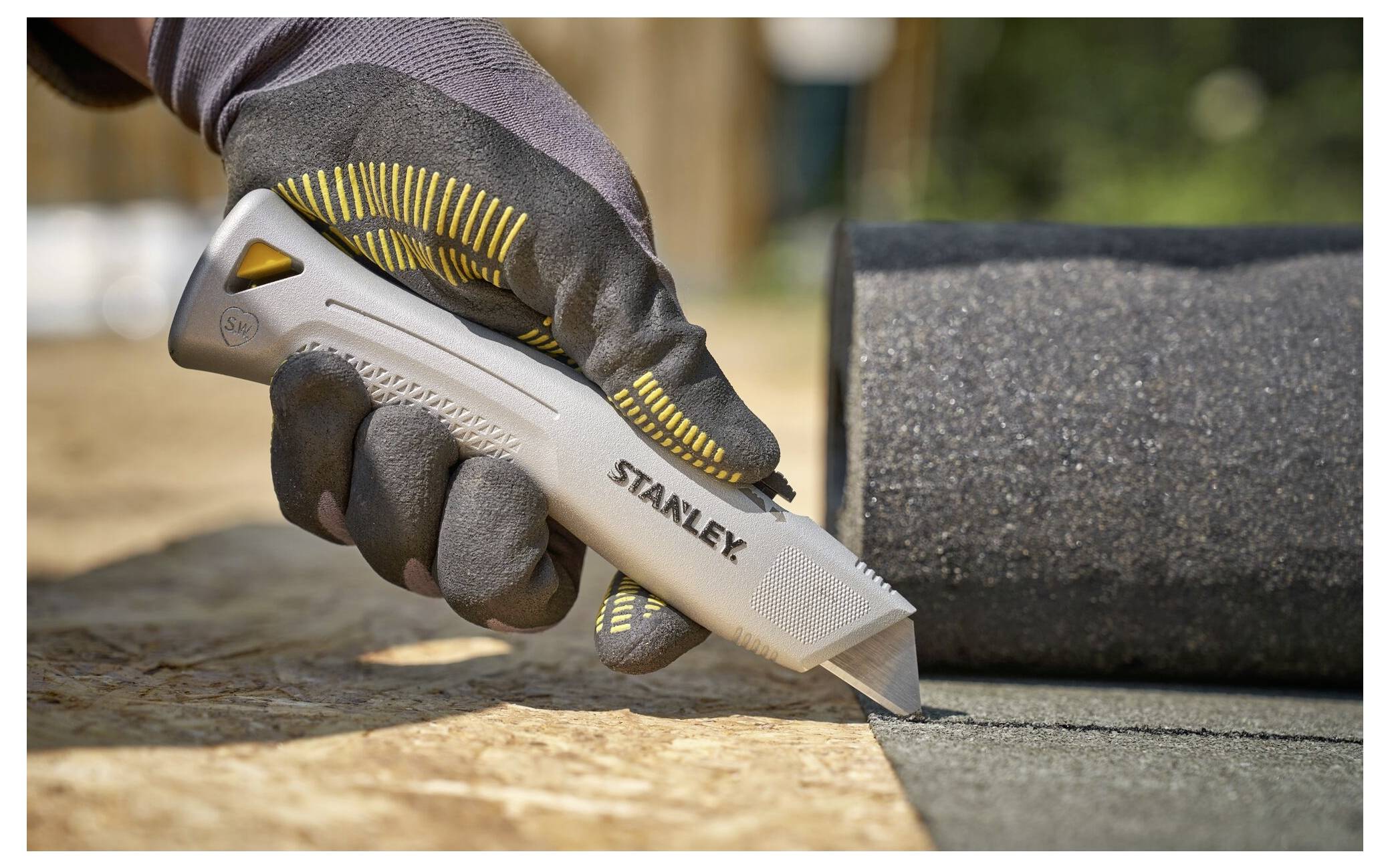 Eine Hand mit einem Handschuh verwendet ein Stanley-Universalmesser, um Dachdeckermaterial auf einer Holzoberfläche zu schneiden, wobei Präzision und Funktionalität hervorgehoben werden.
