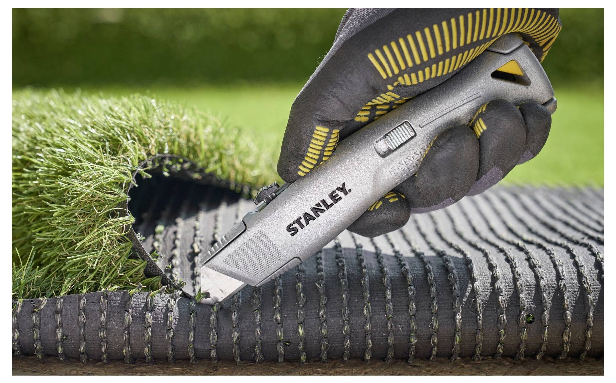 Eine behandschuhte Hand verwendet ein Stanley-Universalmesser zum Schneiden von Kunstrasen und zeigt präzises Trimmen auf einer schwarzen Netzunterlage in einer Gartenumgebung.