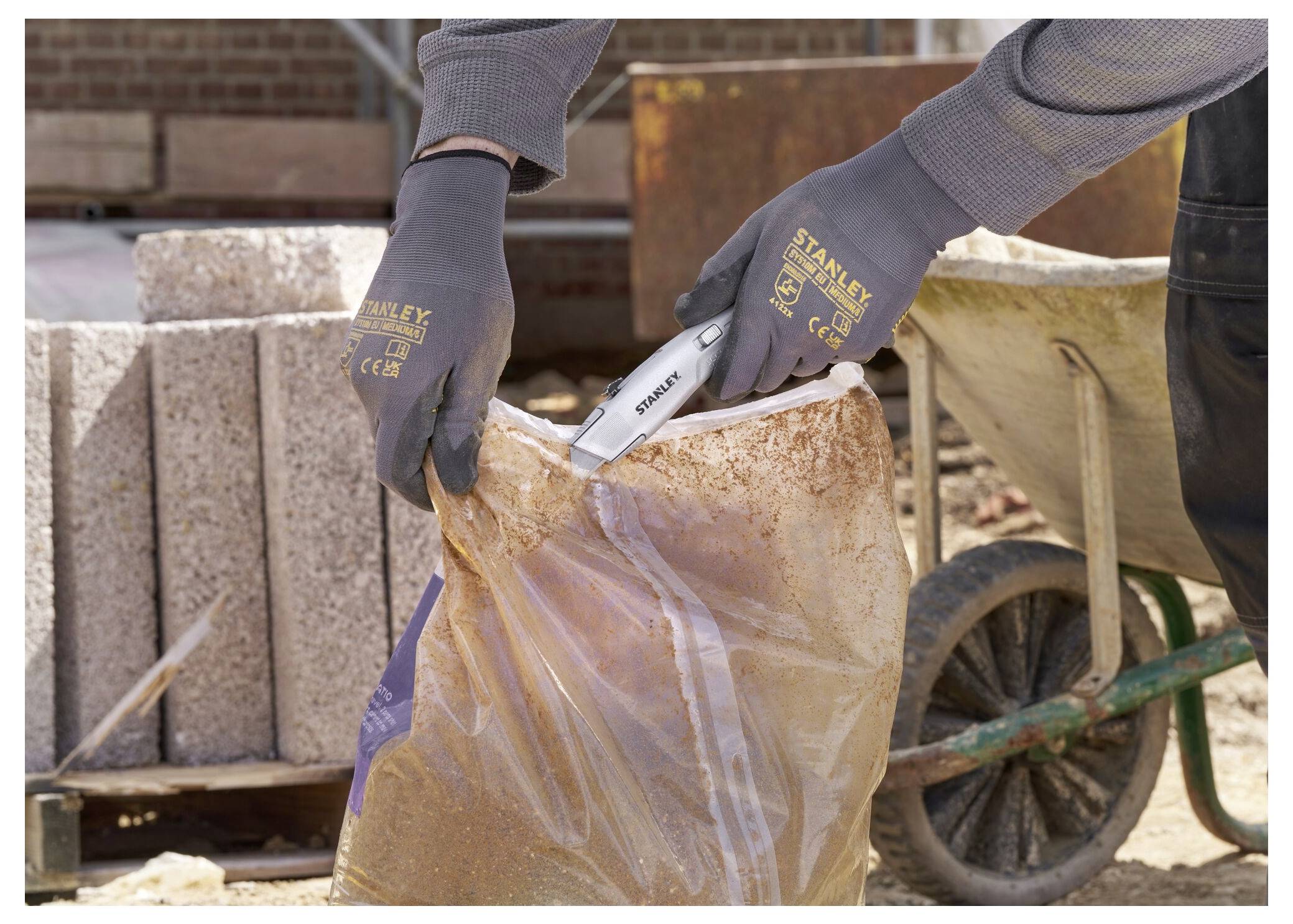 Eine Person mit Handschuhen benutzt ein Stanley-Messer, um einen Plastikbeutel mit Baumaterial zu öffnen, wobei Betonblöcke und eine Schubkarre in der Nähe sind.