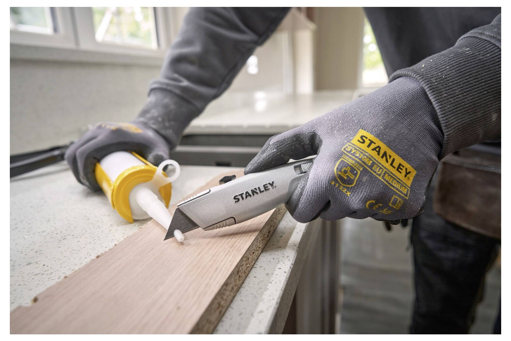 Eine Person, die Stanley-Handschuhe trägt, benutzt ein Stanley-Universalmesser, um die Spitze einer Dichtmassekartucke auf einer Arbeitsplatte zu schneiden.