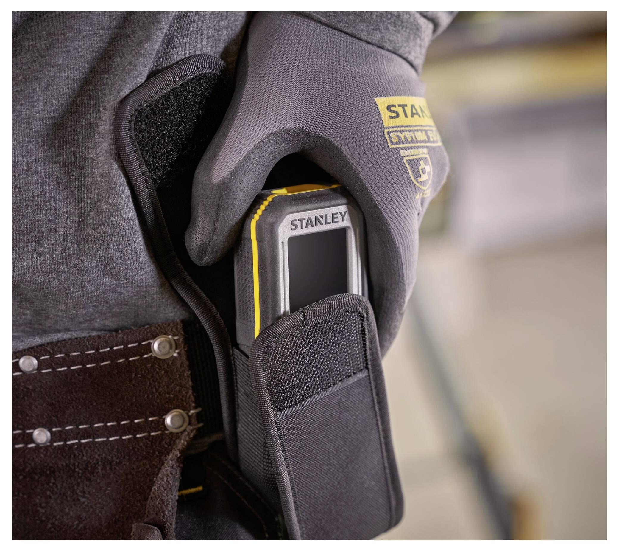 Eine Person, die Handschuhe trägt, legt ein Stanley-Messinstrument in eine Gürteltasche, was Bereitschaft für Außen- oder Bauarbeiten anzeigt.