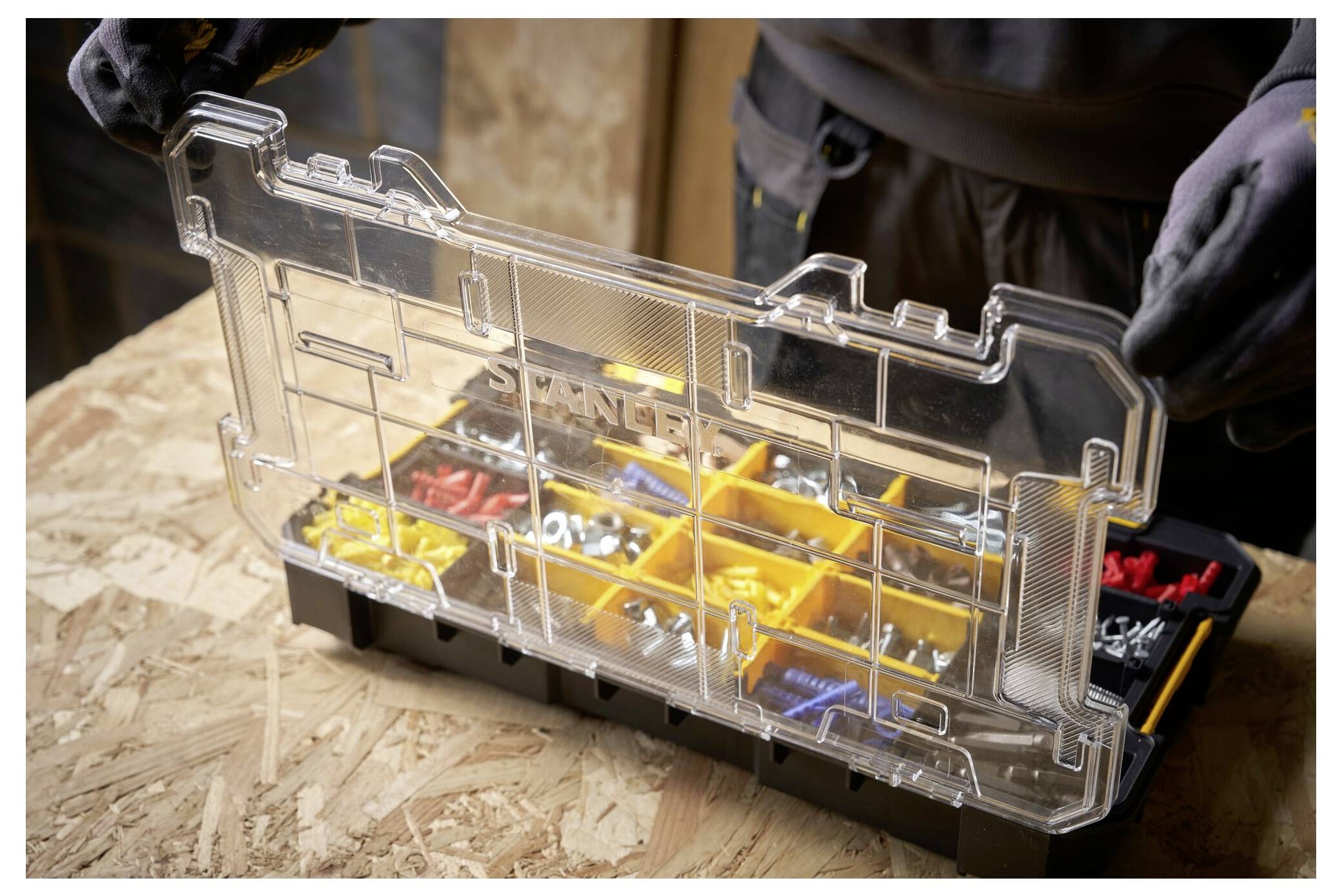 Eine Person mit Handschuhen hält eine offene unterteilte Werkzeugkiste voller verschiedener Schrauben, Bolzen und Dübel auf einer Holzoberfläche.