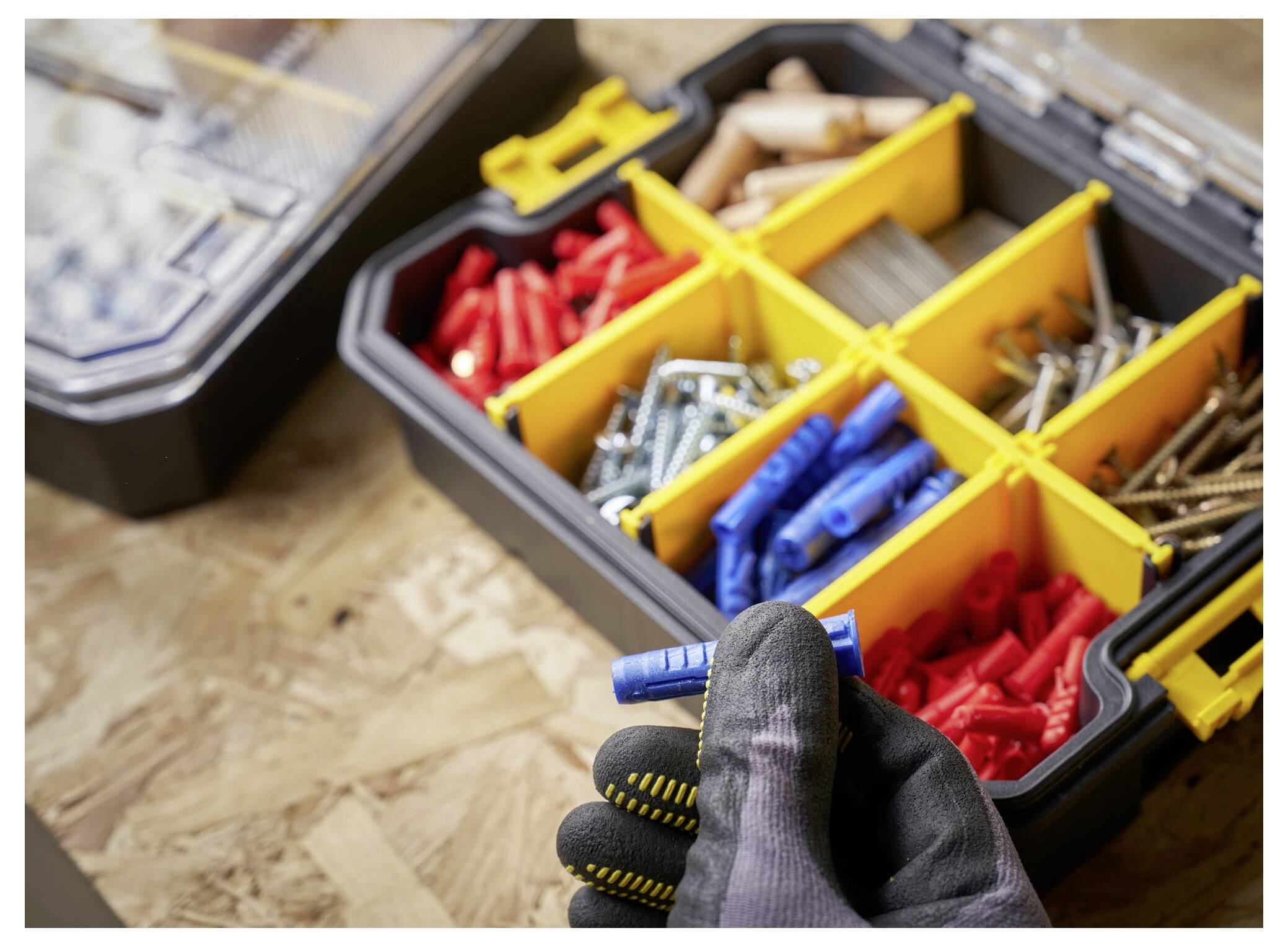 Eine behandschuhte Hand klipst zwei schwarz-gelbe Lagerkästen zusammen, wobei rote Gegenstände in einem Fach sichtbar sind.