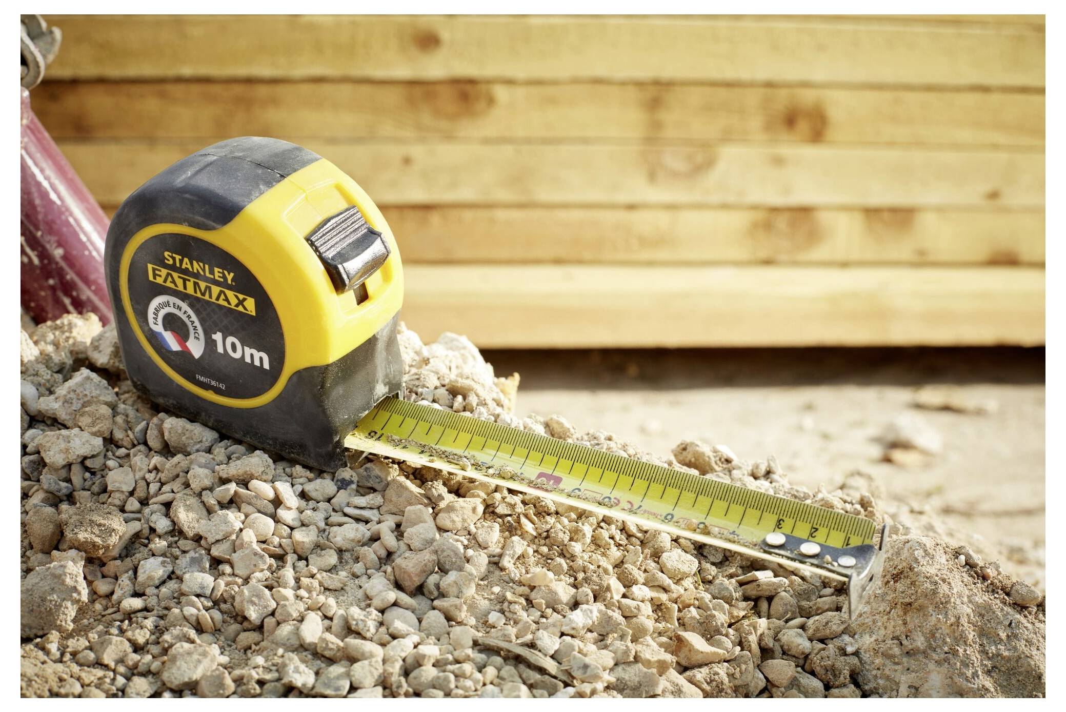 Ein Messstab, der teilweise auf einem Kiesaufschüttung auf einer Baustelle ausgerollt ist, mit hölzernen Bohlen im Hintergrund.