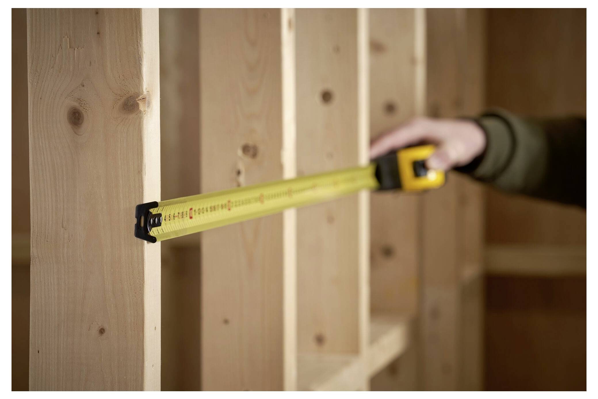 Eine Person misst Holzbalken mit einem gelben Stahllineal und richtet dieses zwischen vertikalen Stützen aus, was auf Bau- oder Schreinarbeiten hindeutet.