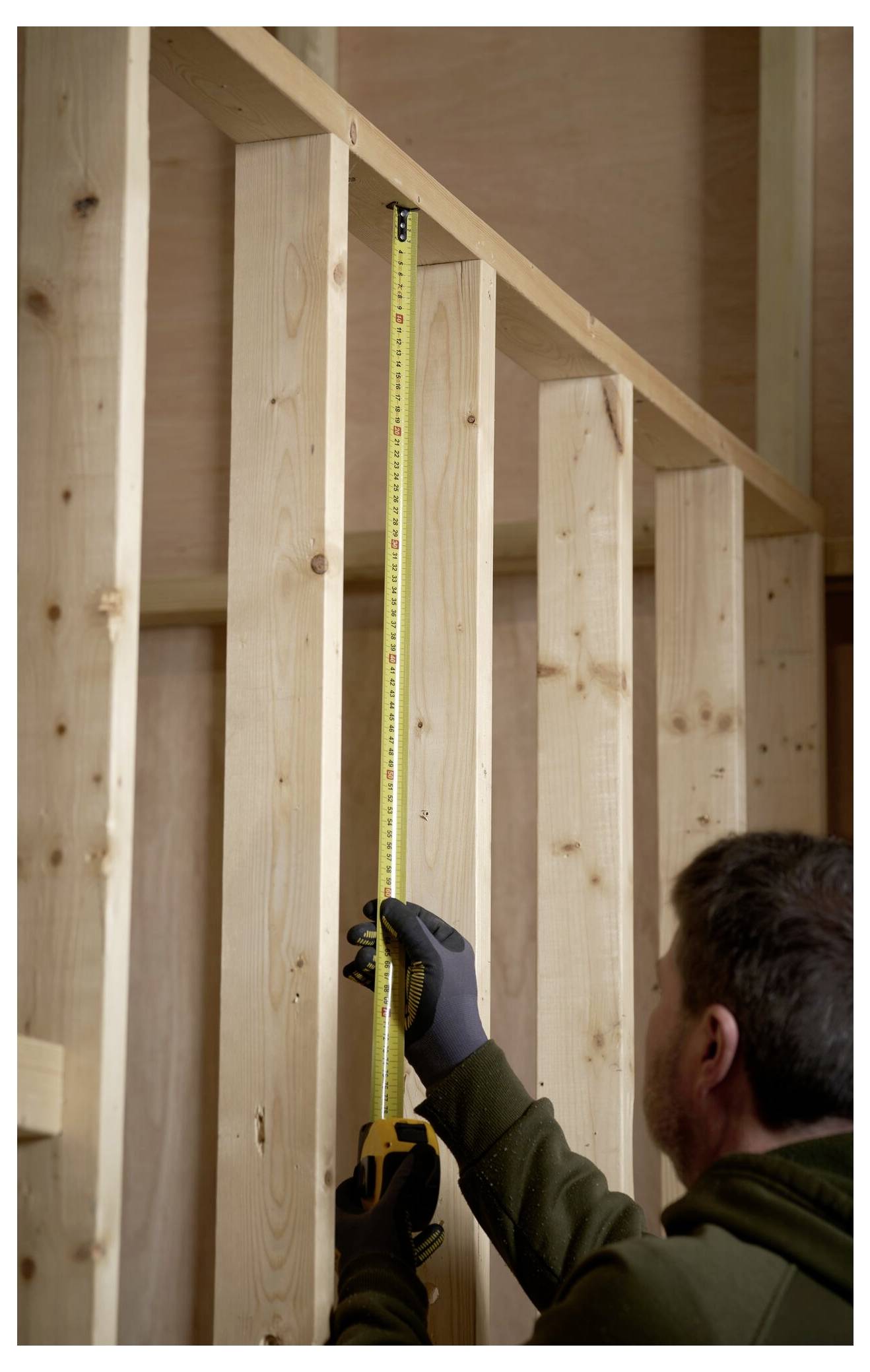 Eine Person, die Handschuhe trägt, verwendet ein Stahllineal auf einem Holzrahmen und arbeitet wahrscheinlich an Konstruktions- oder Zimmermannsarbeiten in einem Gebäude.