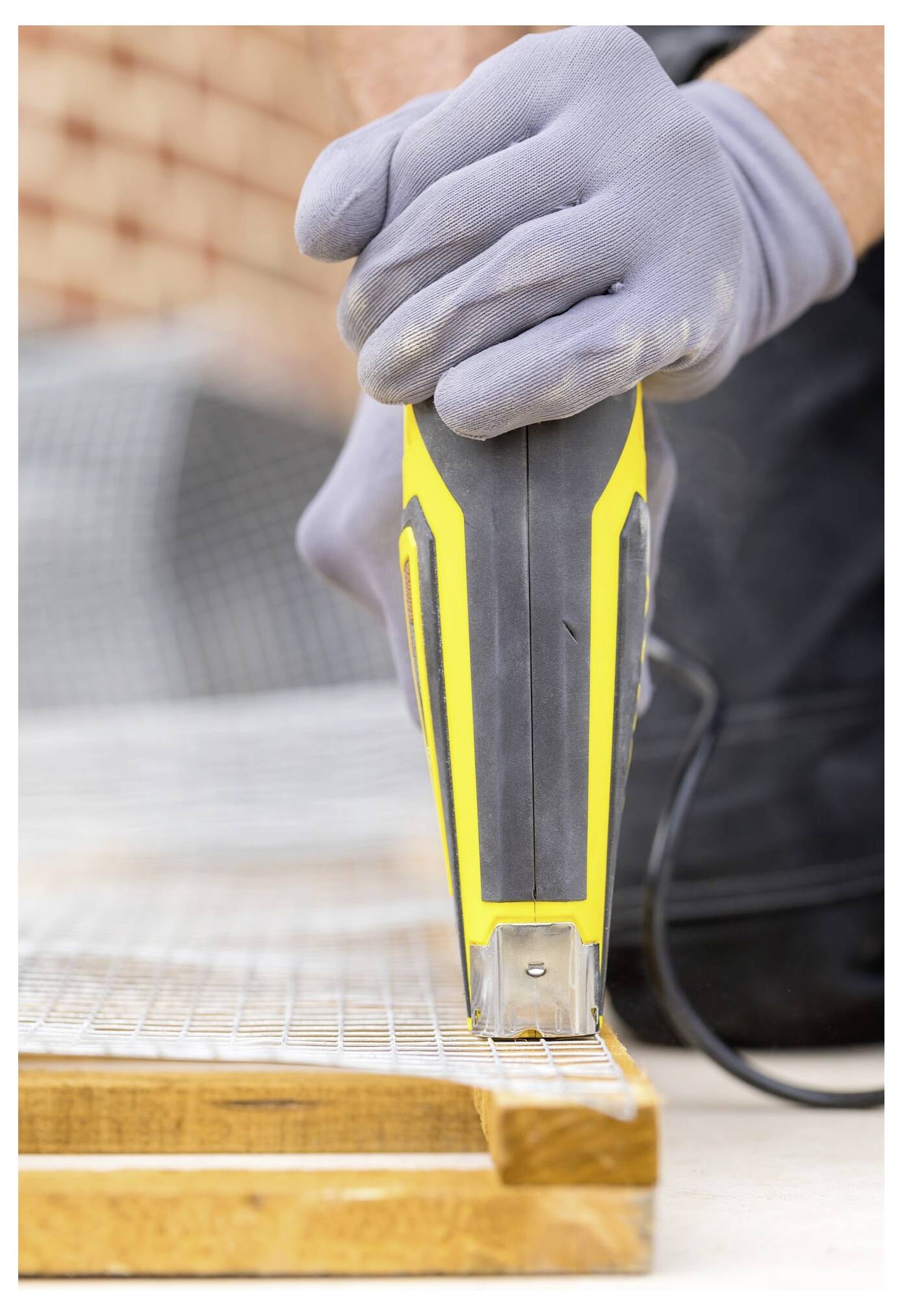Eine Person mit Handschuhen benutzt eine gelb-schwarze Nagelschussmaschine, um ein Drahtgeflecht an einem Holzrahmen zu befestigen, vor einem unscharfen Hintergrund.
