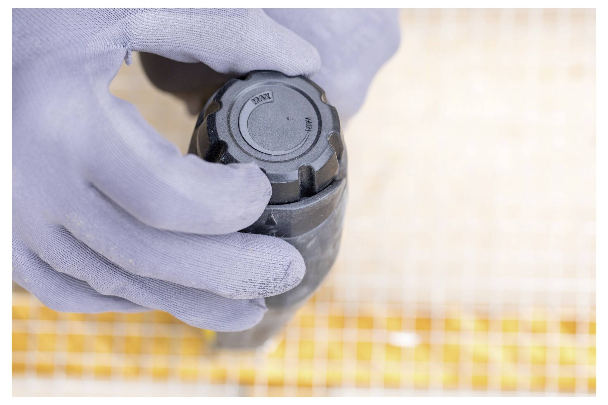 Eine Person, die hellblaue Handschuhe trägt, hält ein schwarzes zylindrisches Objekt, das über einer Oberfläche mit Rastermuster positioniert ist.